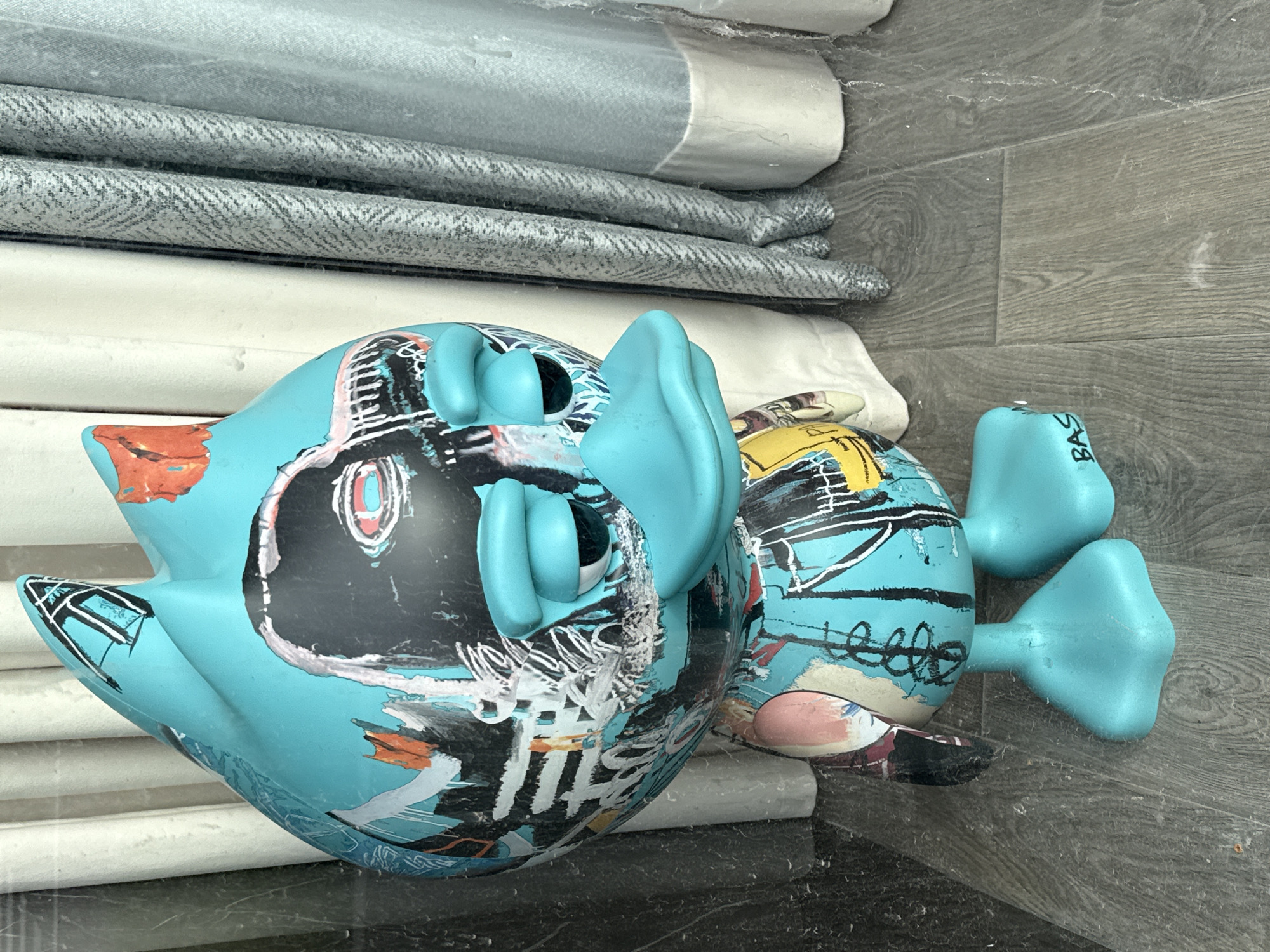 An indoor, eye-level shot captures a vibrant turquoise art figure, designed in a stylized, cartoonish manner, lying on its side. The figure's surface is adorned with abstract, graffiti-like artwork in black, white, red, orange, and yellow hues, reminiscent of Jean-Michel Basquiat's style. It features prominent, round eyes, small facial features, and paddle-like hands and feet, all in the same bright turquoise. On one of the feet, the partial text "BAS" is visible. Another similar, though less visible, figure lies partly behind it.
The setting appears to be a storage or display area, possibly a shelf or cabinet. Above the figures, a stack of folded textiles is visible. These include a light-colored, possibly white or light grey, fabric at the top, followed by two pieces of grey fabric with a distinct herringbone pattern. Below these, another plain white textile is stacked. The surface directly beneath the figures is dark and subtly reflective, possibly stone or a dark laminate. To the right, a vertical surface with grey wood-grain texture is visible, suggesting a wall panel or flooring. The lighting indicates an indoor environment. The scene is set in Moscow, Russia.
