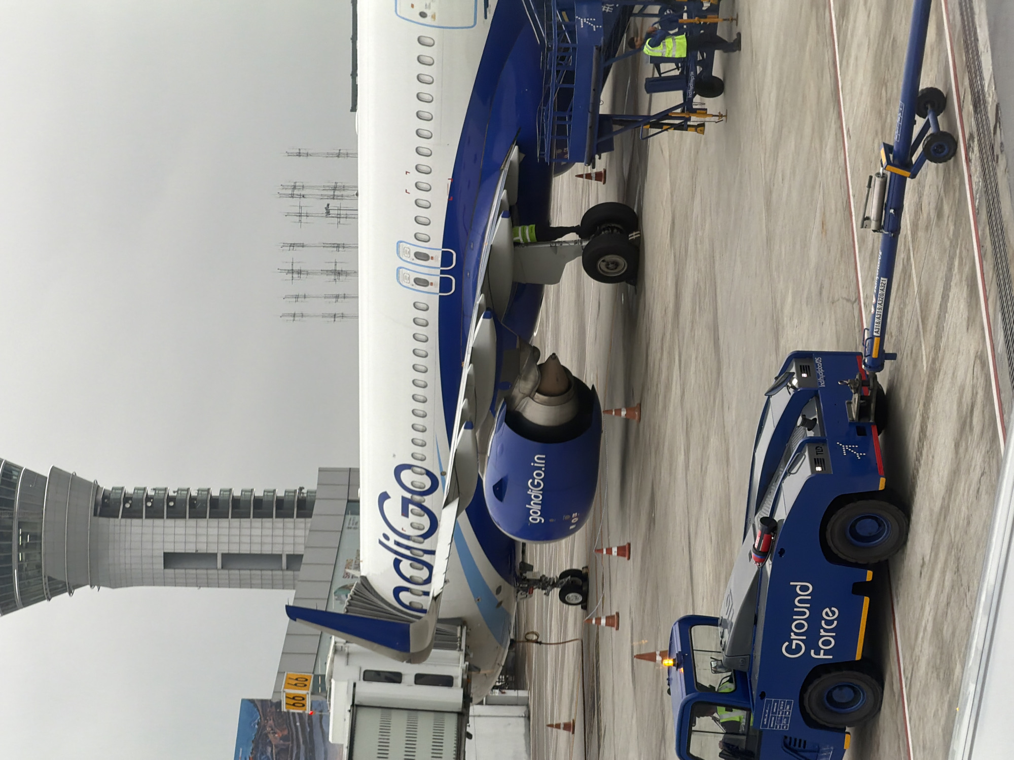 An Indigo Airlines airplane is parked at the Rajiv Gandhi International Airport in Hyderabad, India. A blue "Ground Force" tow vehicle is attached to the aircraft's front landing gear, suggesting it is being prepared for pushback or towing. A ground crew member in a high-visibility vest is visible near the tow vehicle, and another is on a boarding ramp next to the plane. In the background, the airport's control tower is prominent. The scene is set on a tarmac under an overcast sky, with various airport equipment and safety cones present.