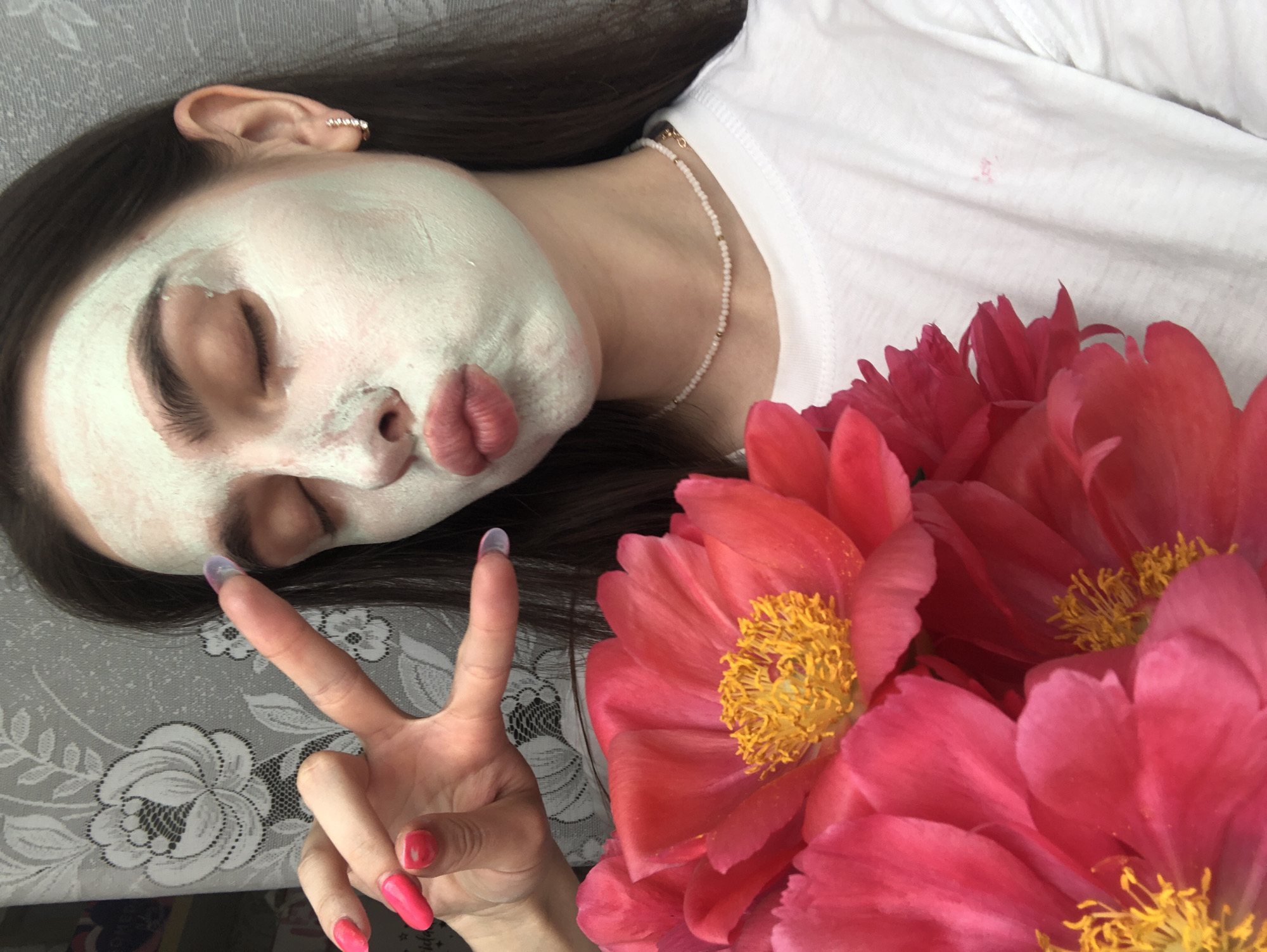 A young woman with dark hair and a white t-shirt is holding a bouquet of pink peonies while applying a green face mask. She has her eyes closed and her lips pursed, giving a kiss. Her right hand is raised, with her index and middle fingers forming a peace sign. The background is a floral wallpaper. The location is not specified in the image.