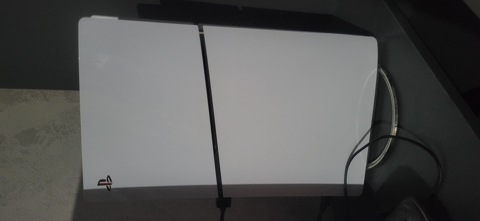A white Playstation 4 console is sitting on a dark surface, likely a shelf or entertainment center, located indoors. The console is a two-piece design separated by a black vertical line. Wires are visible near the right side of the console. The wall next to the Playstation has a textured pattern. The location is Unknown, Unknown.