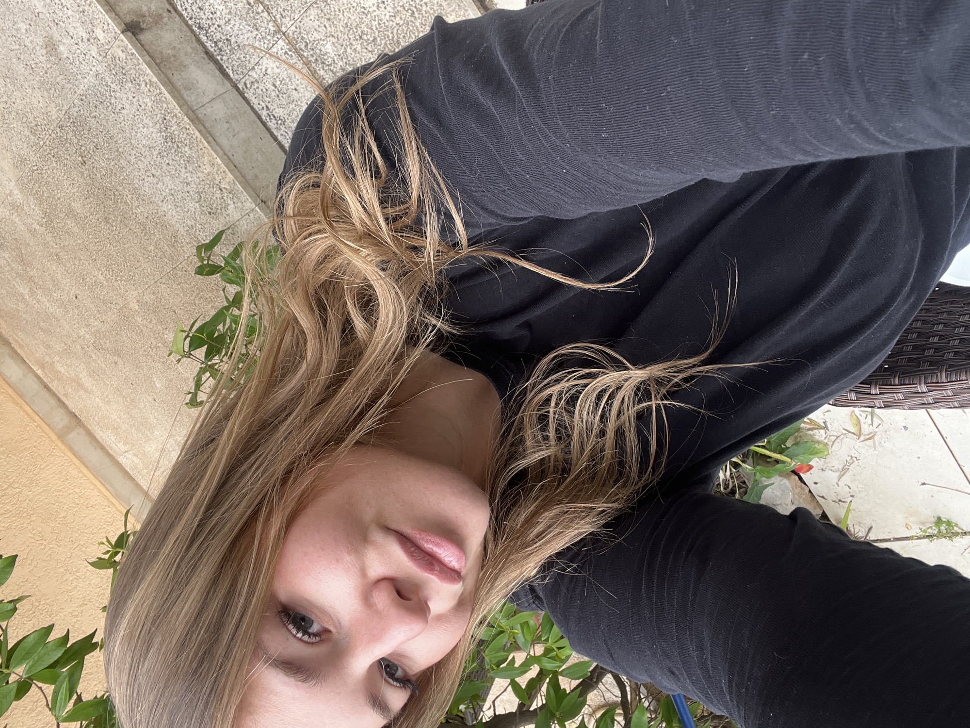 A medium shot captures a woman from a slightly downward angle, with her face occupying the lower left portion of the frame. She has fair skin, light brown hair, and appears to be wearing a black long-sleeved shirt.
The backdrop features a concrete staircase in the upper left, transitioning to outdoor greenery and a light-colored wall on the left. To the right, vegetation and possibly outdoor furniture are visible, suggesting an outdoor setting.
Based on the image's origin information, the location is Krimovice, Montenegro. No specific objects or activities suggest further details about the context of the scene.