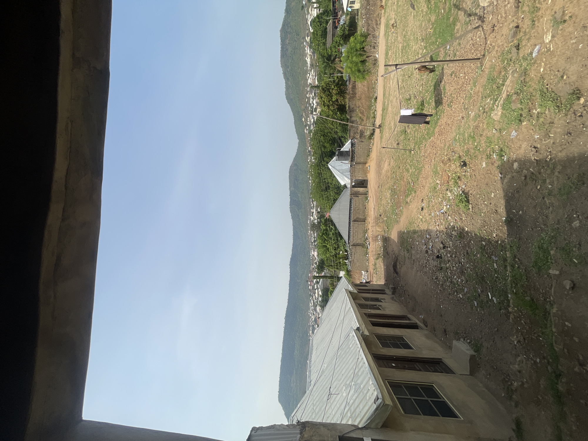 This image captures a wide outdoor scene from what appears to be an elevated position, possibly looking out from a window or balcony of a concrete building, whose dark frame partially obscures the left side of the view. The overall orientation of the image is vertical, with the sky to the left and ground to the right, but it's best understood if rotated 90 degrees counter-clockwise.
The setting is a residential area at the base of a range of lush, green mountains, indicative of the natural landscape around Lokoja, Nigeria. The sky is clear and bright blue, suggesting daytime, likely morning or afternoon given the illumination.
In the mid-ground, there are several simple, unpainted concrete buildings, many with corrugated metal roofs, reflecting the sunlight. These structures are scattered among patches of dry, reddish-brown dirt, indicating unpaved roads and yards, and some sparse green vegetation. Power lines are visible, strung between poles, hinting at basic infrastructure.
A notable activity in the scene is laundry drying on a clothesline, strung between wooden poles in an open, grassy-dirt area. A dark garment and a lighter one are hanging. This area also shows signs of general refuse, with scattered plastic bags and other debris on the ground. Further into the mid-ground, a lone dog can be seen sitting on the dirt.
The background is dominated by the imposing, tree-covered mountains, which stretch across the horizon. More small, white or light-colored buildings dot the lower slopes of these mountains, extending the residential settlement. No specific individuals or discernible text are visible in the image. The overall impression is one of a functional, semi-rural community nestled within a mountainous landscape.