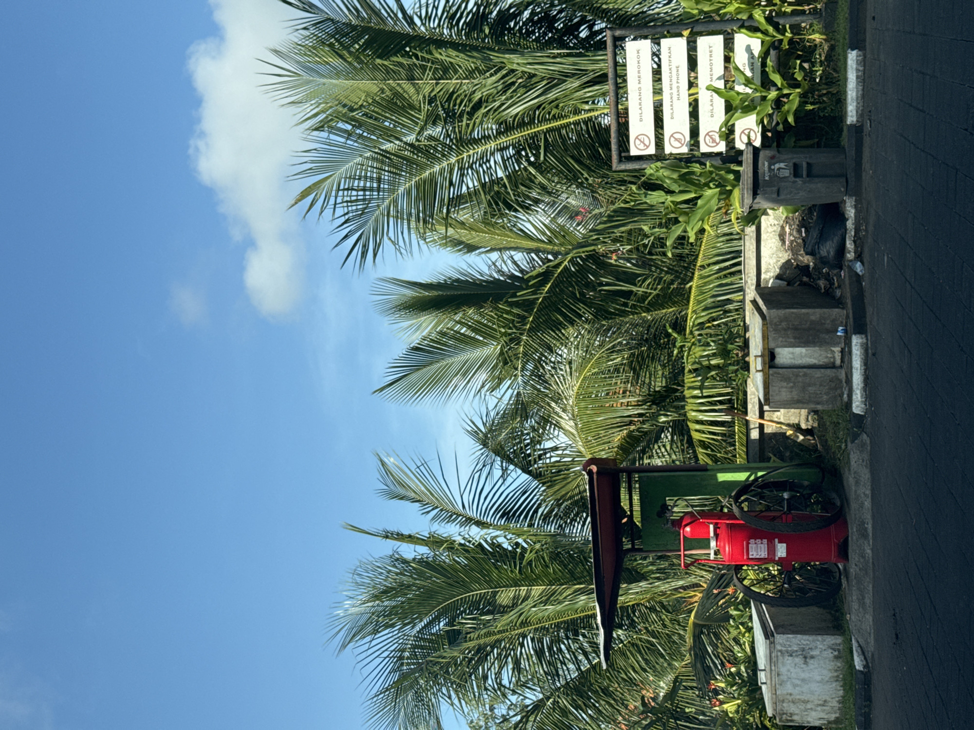 The image captures a scene in Ungasan, Indonesia, focusing on a roadside setup. On the right edge is a paved road. Adjacent to the road is a concrete structure. Next to the concrete structure is a green fire extinguisher cart. Behind the cart is lush tropical vegetation, primarily palm trees. A signpost is positioned among the palm trees, with several signs displaying prohibitions. The sky is visible in the upper left corner of the image.