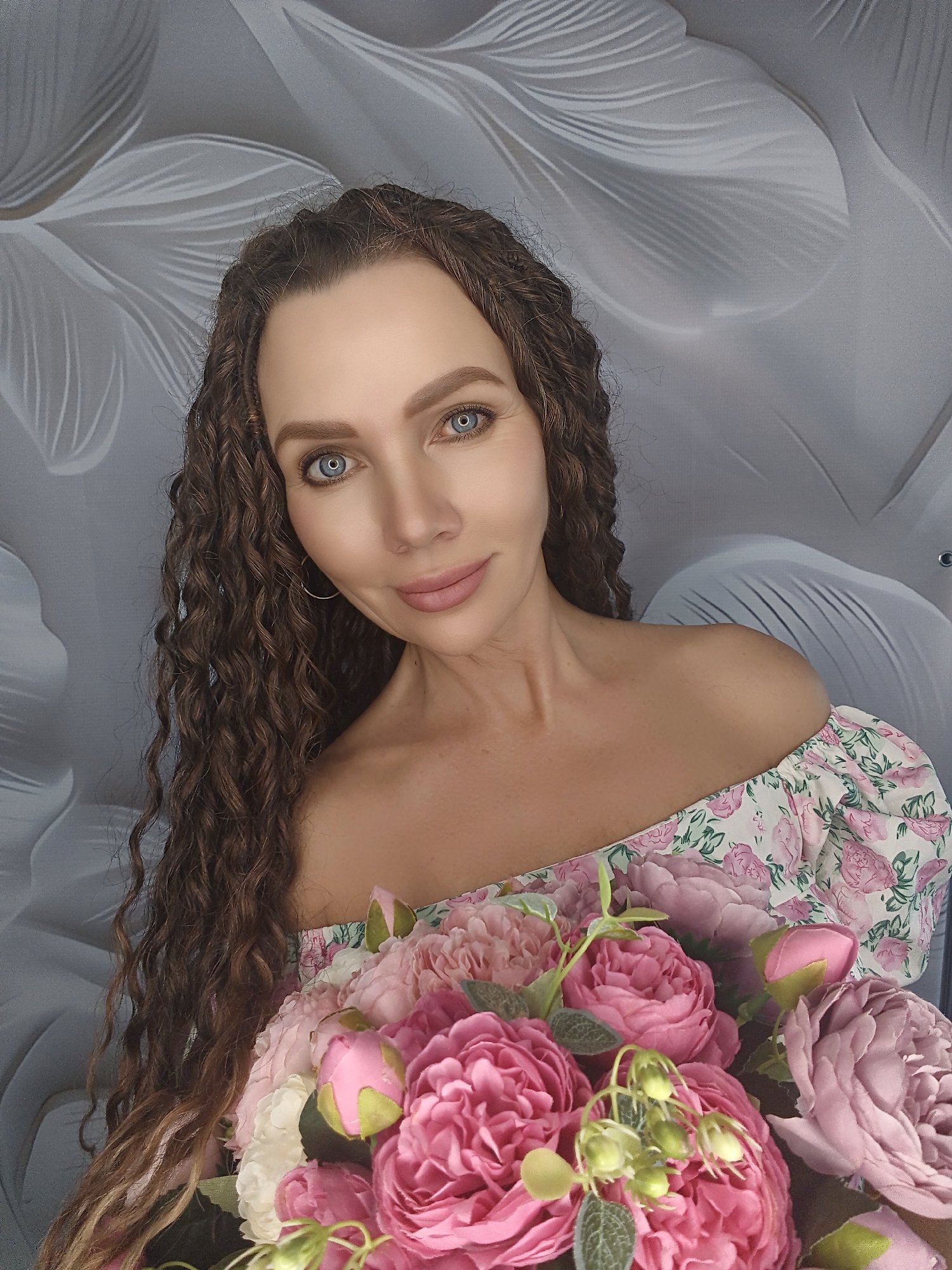 A close-up, waist-up, studio portrait shows a woman with long, curly brown hair and blue eyes. She is wearing an off-the-shoulder floral dress and holding a bouquet of pink and white flowers. The background is a grey, textured wall with a light-colored floral pattern. The lighting is soft and diffused, creating a gentle ambiance. The woman is looking directly at the camera with a slight smile. The overall image conveys a sense of beauty and femininity.