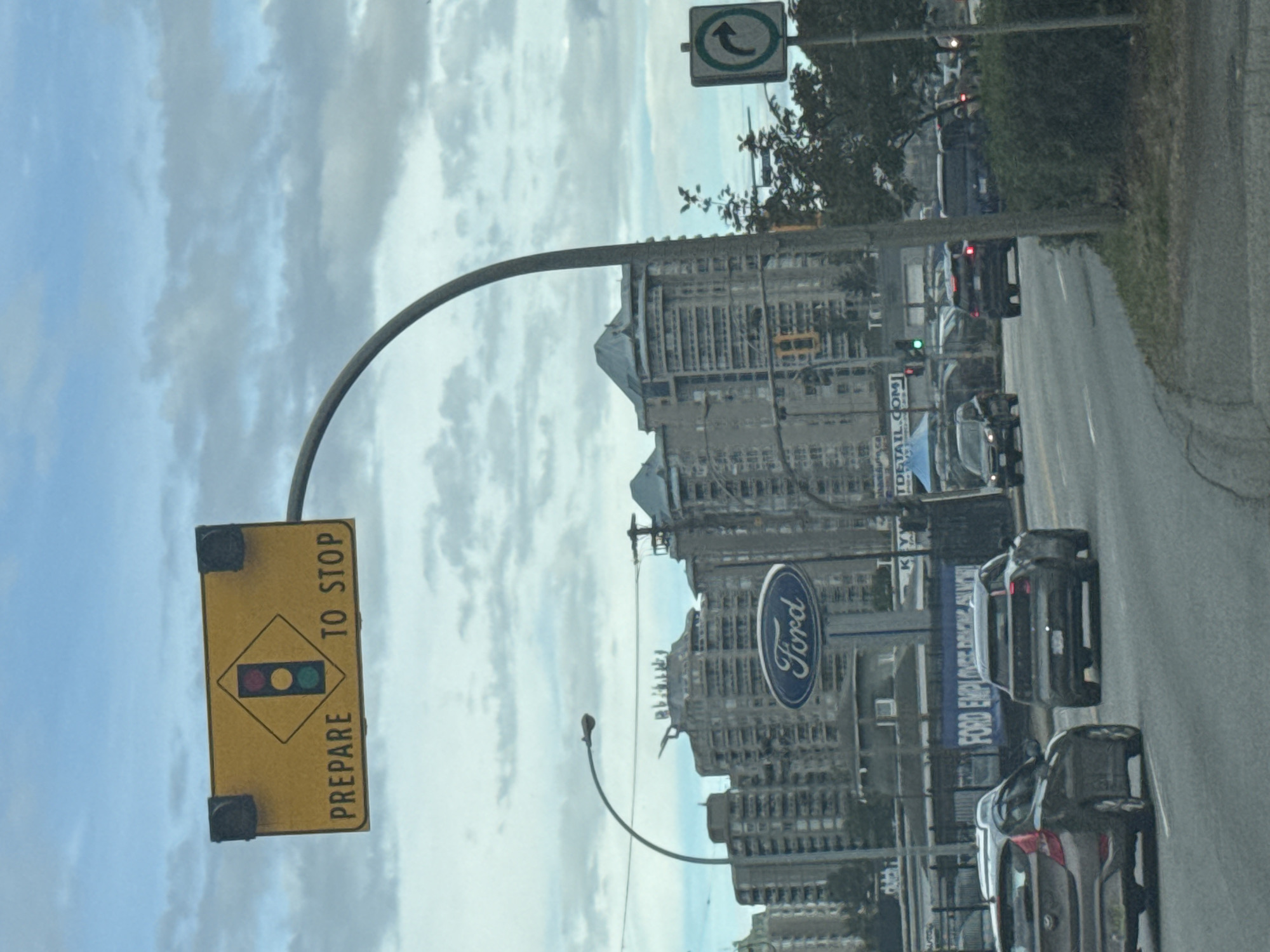 An overcast day in New Westminster, Canada, is depicted in this street-level photograph. The primary subject is a yellow diamond-shaped traffic sign with a traffic light symbol and the words "PREPARE TO STOP" below it, mounted on a curved metal pole. The traffic light in the symbol shows a green light illuminated.
In the background, several multi-story apartment buildings with a modern architectural style dominate the skyline. A large "Ford" dealership sign is visible, suggesting a commercial area. The road in the foreground is asphalt with lane markings. Several vehicles are on the road, including a dark-colored sedan in the right lane and a brown hatchback in the left lane, both moving forward. Further down the road, other vehicles are visible, including a dark SUV and a blue SUV. A traffic light displaying a green signal is visible on the right side of the road. A directional sign with a right-turn arrow is also present on the right. The scene is consistent with a suburban or urban street environment in New Westminster, likely near a commercial district.