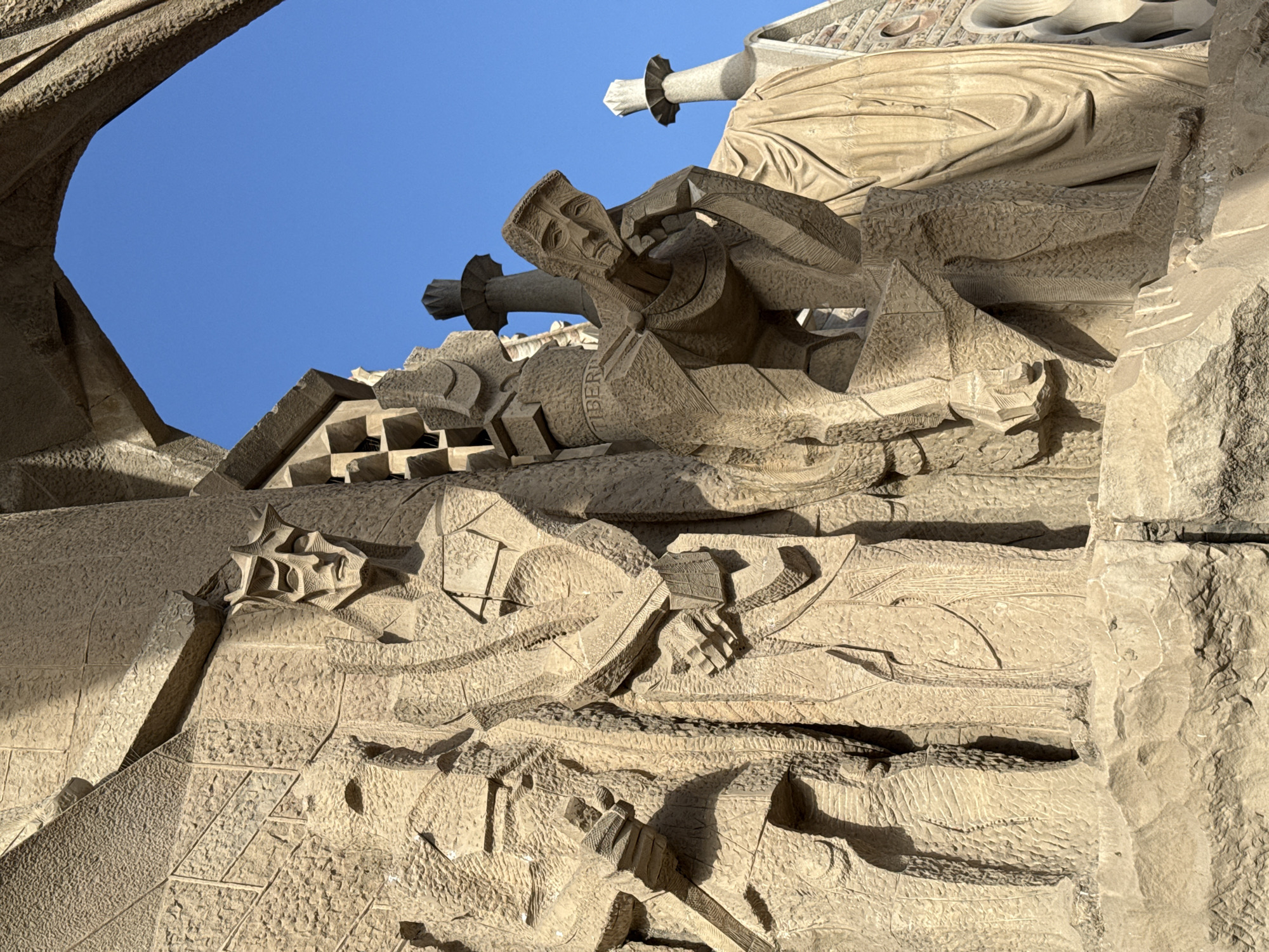 The image depicts a close-up view of a stone relief sculpture on the exterior of the Sagrada Familia in Barcelona, Spain. The sculpture features several figures carved in a stylized, almost cubist manner. In the foreground, on the right, a figure is depicted kneeling, with its head bowed and hands clasped, conveying a sense of sorrow or contemplation. To the left of this figure, a taller, standing figure is shown, also with a somber expression, holding what appears to be a scroll or tablet. The stonework is highly textured, with rough surfaces and deep carvings that create strong shadows. Above and behind the figures, more architectural elements of the Sagrada Familia are visible, including intricate patterns and arched openings. The sky, a vibrant blue, is visible through one of these openings. The overall impression is one of solemnity and intricate craftsmanship.
