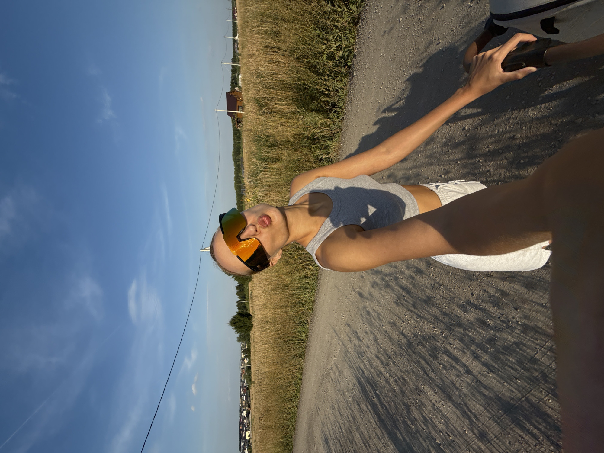 A woman wearing sunglasses and a grey crop top is taking a selfie while standing on a dirt road in a rural area, likely on the outskirts of Podshivalovo, Russia. She is puckering her lips for the camera. Behind her, a field of dry grass stretches towards a treeline and a few houses are visible in the distance. The sky is blue with some wispy clouds and power lines run across the frame. The sun casts long shadows on the road. The woman is holding onto handlebars of what appears to be a bicycle.