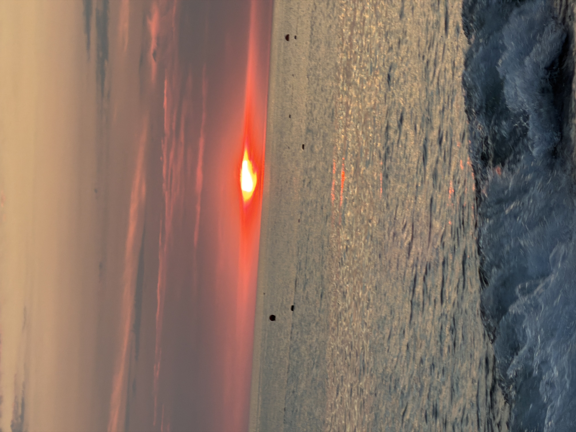 A sunset over the Black Sea in Sochi, Russia, is depicted in this photograph. The sky is painted with hues of orange, pink, and yellow, with thin, wispy clouds stretching across the horizon. The sun, a brilliant red orb, is low in the sky, casting a warm glow on the water's surface. The sea is calm, with gentle waves lapping at the shore. In the distance, several buoys are visible, marking a section of the water. The foreground is dominated by a breaking wave, its white foam contrasting with the darker blue of the ocean.