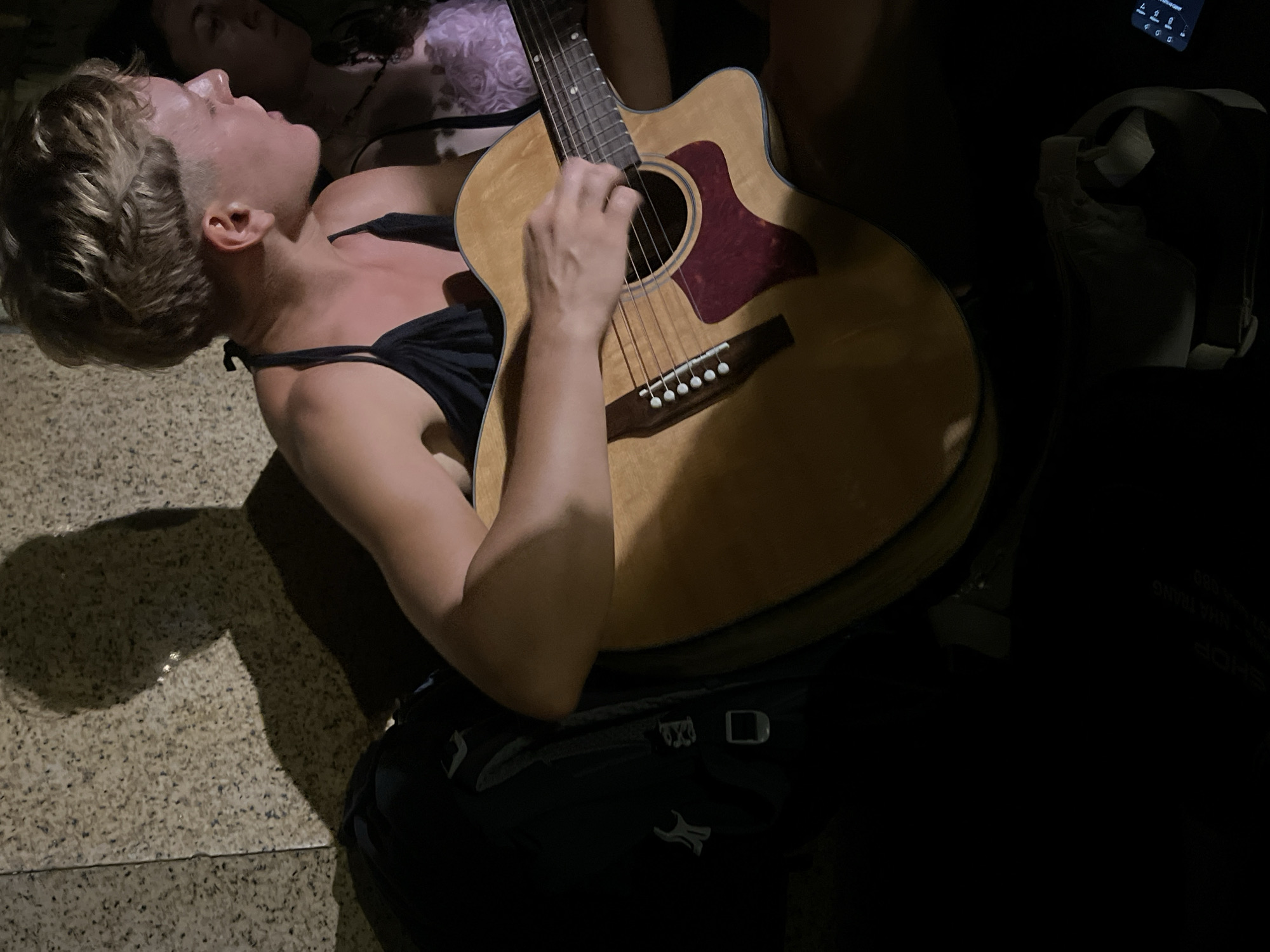 A person with short blond hair, wearing a black top with thin straps, is playing an acoustic guitar. The guitar has a reddish-brown pickguard and light-colored wood. A person is lying behind them, partially visible. The person playing the guitar has a backpack underneath the guitar. The surface is a patterned stone. In the upper right is the edge of a white object and a dark object with illuminated symbols on it. The location is Nha Trang Ward, Vietnam.
