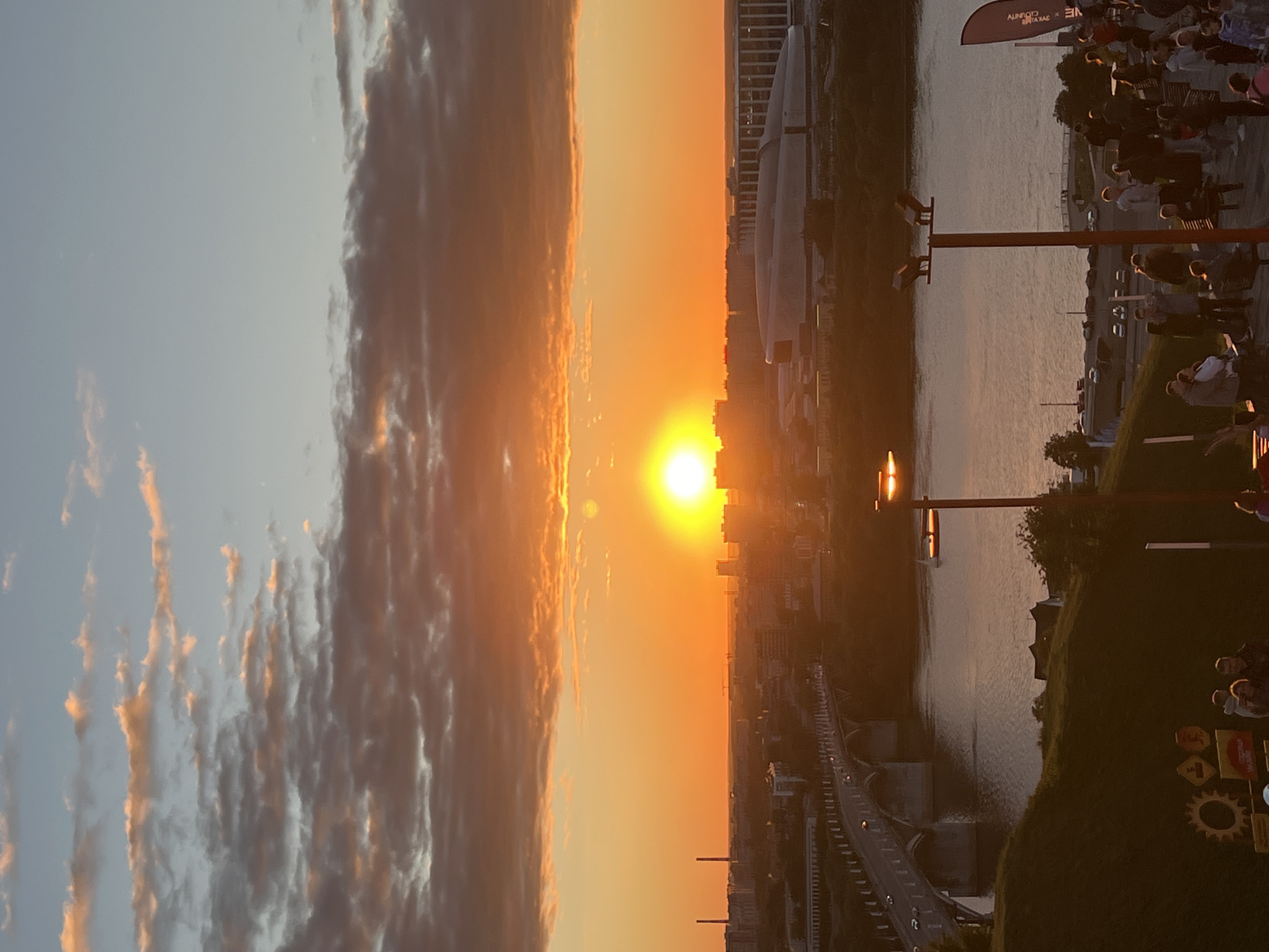 The image captures a vibrant sunset over the city of Nizhny Novgorod, Russia, viewed from the Upper Volga Embankment. The sun, a brilliant orb of orange, is positioned low on the horizon, casting a warm glow across the sky and illuminating the city skyline. The sky is a canvas of warm hues, with shades of orange, yellow, and hints of pink blending into the blue above. Clouds, tinged with the sunset's light, add texture and depth to the celestial scene.
In the foreground, a section of the embankment is visible, with a pathway leading down towards the Volga River. Several streetlights, their lamps glowing warmly, are strategically placed along the embankment, illuminating the area for evening visitors. A group of people are gathered on the embankment, some standing and conversing, while others are seated, enjoying the view. They appear to be engaged in leisurely activities, their silhouettes softened by the ambient light.
Across the river, the Nizhny Novgorod Stadium, a modern architectural marvel, stands prominently on the opposite bank. Its distinctive, futuristic design is partially visible, its form contrasting with the older structures of the city. A bridge, likely the Kanavinsky Bridge, arches gracefully over the Volga River, with vehicles traversing its lanes, their headlights creating streaks of light in the fading daylight. The river itself reflects the warm tones of the sunset, creating a shimmering expanse of water.
Further into the city, buildings of varying architectural styles dot the landscape, interspersed with glimpses of industrial structures and green spaces. The overall impression is one of urban life coexisting harmoniously with the natural beauty of the sunset and the river. The scene evokes a sense of tranquility and the end of a day, with the city bathed in the golden light of the setting sun.