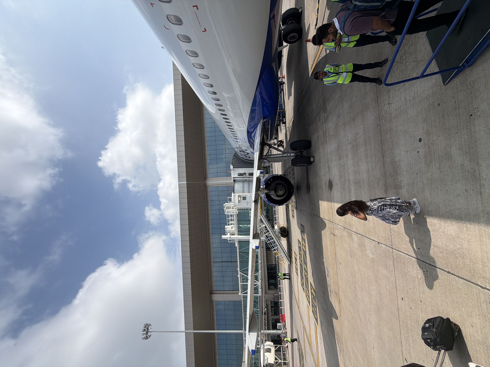 An aerial view shows an Indigo Airlines airplane parked at the terminal in Vikram Nagar, India, under a cloudy blue sky. The aircraft, with its distinctive blue and white livery, is connected to the terminal via a jet bridge. Several people are visible on the tarmac, including passengers and airline staff in high-visibility vests. One woman in a patterned dress stands on the tarmac, while a suitcase rests beside her. A staircase leads up to the airplane's entrance, and an orange traffic cone is placed nearby. The airport terminal building features a modern design with large glass windows. The scene is characterized by bright sunlight casting shadows across the concrete tarmac, marked with yellow lines indicating parking positions.
