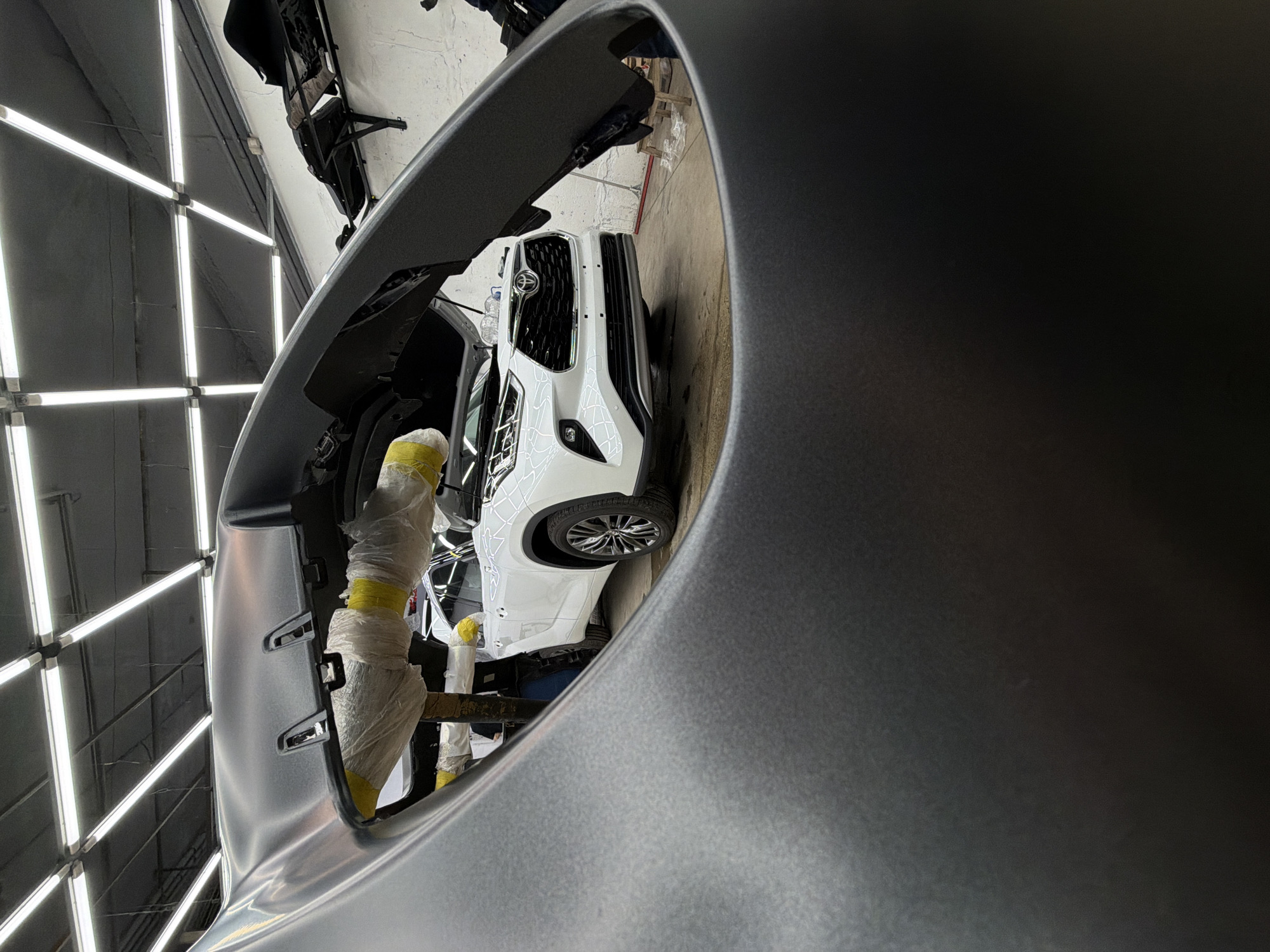 The image depicts a partially disassembled white Toyota SUV inside what appears to be a body shop. The SUV's front end is visible through the partially removed and gray-painted bumper, which is in the foreground. Other car parts are being held together with masking tape. The body shop is brightly lit with fluorescent lights. The location is unknown, and the description provided in the prompt is incorrect.