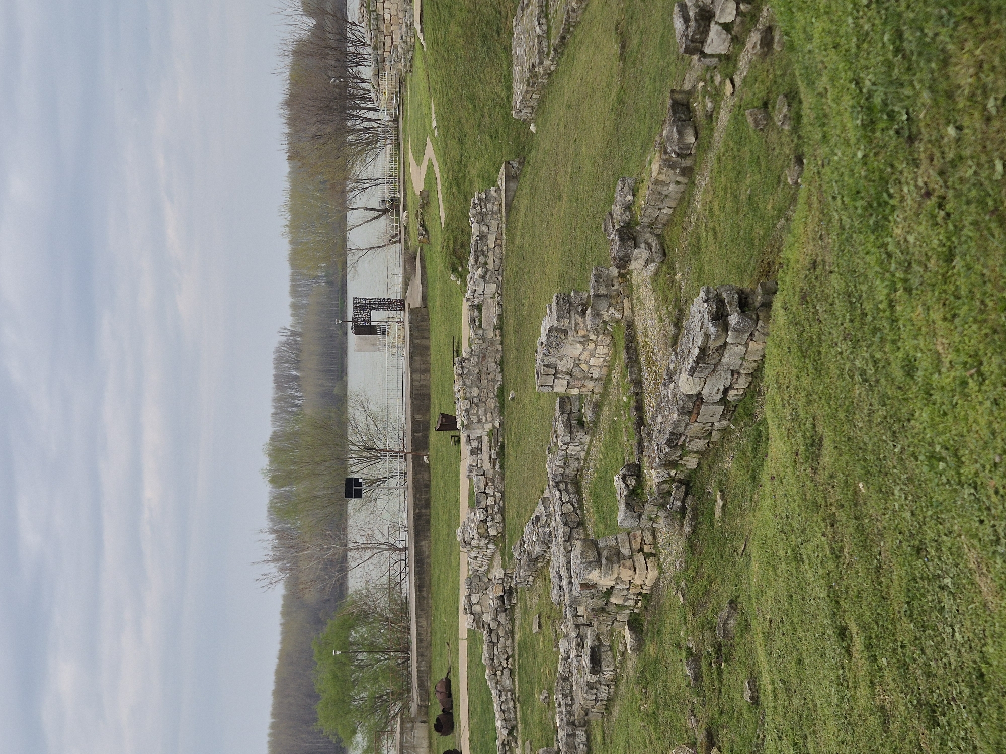 The image shows ruins on a grassy area next to a body of water. The ruins are made of stacked stones. A metal railing runs along the water's edge. There are trees in the background along the water. The sky is overcast. The location appears to be near Silistra, Bulgaria, possibly associated with Roman ruins in the area.