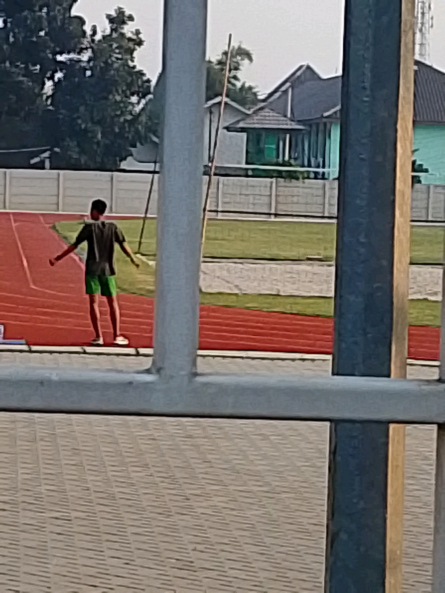 A person stands on a red track, arms outstretched, wearing a dark t-shirt and green shorts. The track appears to be part of a sports facility, with a grassy area and buildings in the background. The scene is viewed through a series of gray metal bars, suggesting the person is within a fenced area. The buildings in the background are colorful, with one having a green roof. The location is identified as Cibinong, Indonesia.
