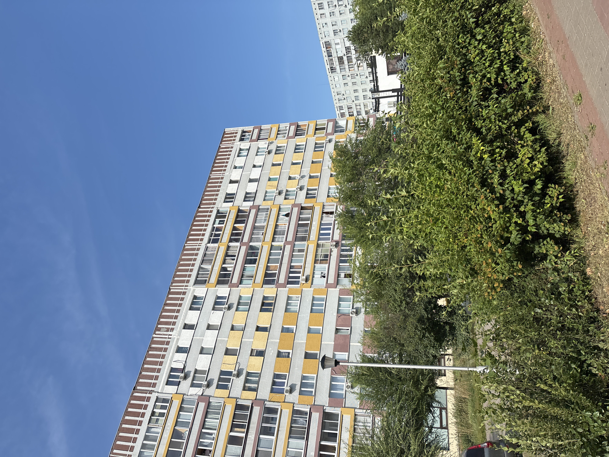 The image shows a residential area in Naberezhnye Chelny, Russia, under a clear blue sky. In the foreground, dense green bushes and trees partially obscure a sidewalk paved with red bricks and a grassy area. Behind the vegetation, a tall, multi-story apartment building with a colorful facade, featuring grey, yellow, and brown sections, is prominently visible. Several windows and air conditioning units are visible on the building. A lamppost stands in front of this building. Further in the background, another, similar apartment building with a predominantly grey facade is visible. The scene appears to be a typical urban residential neighborhood. There are no people or animals in the frame.