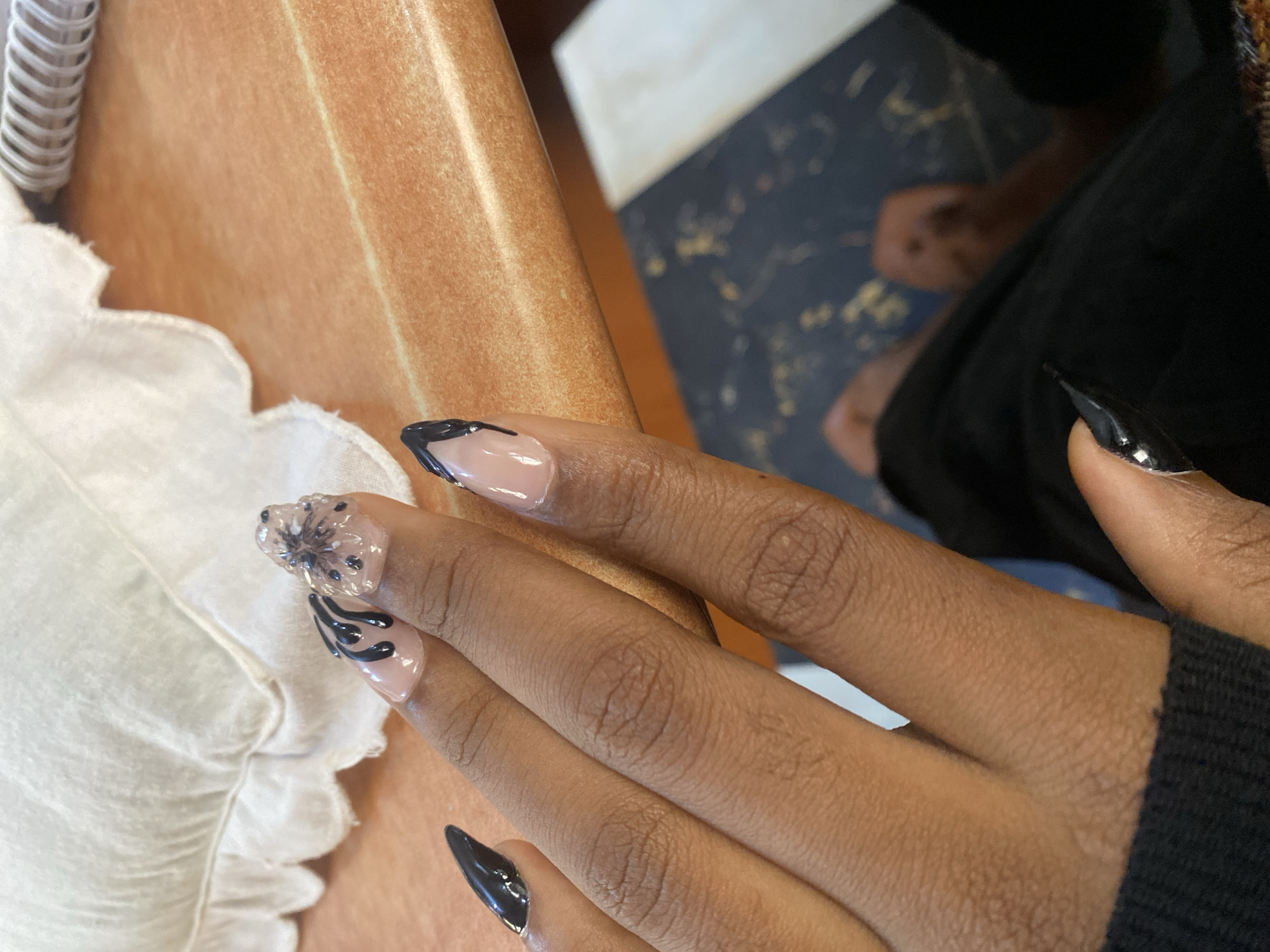 In an indoor setting, a close-up shot focuses on a person's manicured hand resting on a textured, light brown surface, possibly a table or desk. The hand exhibits long, almond-shaped artificial nails. Several nails are adorned with a French manicure design featuring a sheer pink base and black tips. One nail is decorated with black abstract lines and what appears to be a 3D embellishment, possibly a flower or abstract design made of clear material with black speckles. The background shows a white ruffled fabric object on the left and a blurry dark surface with some lighter patterns on the bottom right, suggesting a floor or patterned mat. The overall lighting is bright, with some shadows cast on the skin and surface. The scene does not provide any specific visual cues to determine its location within Jos, Nigeria.