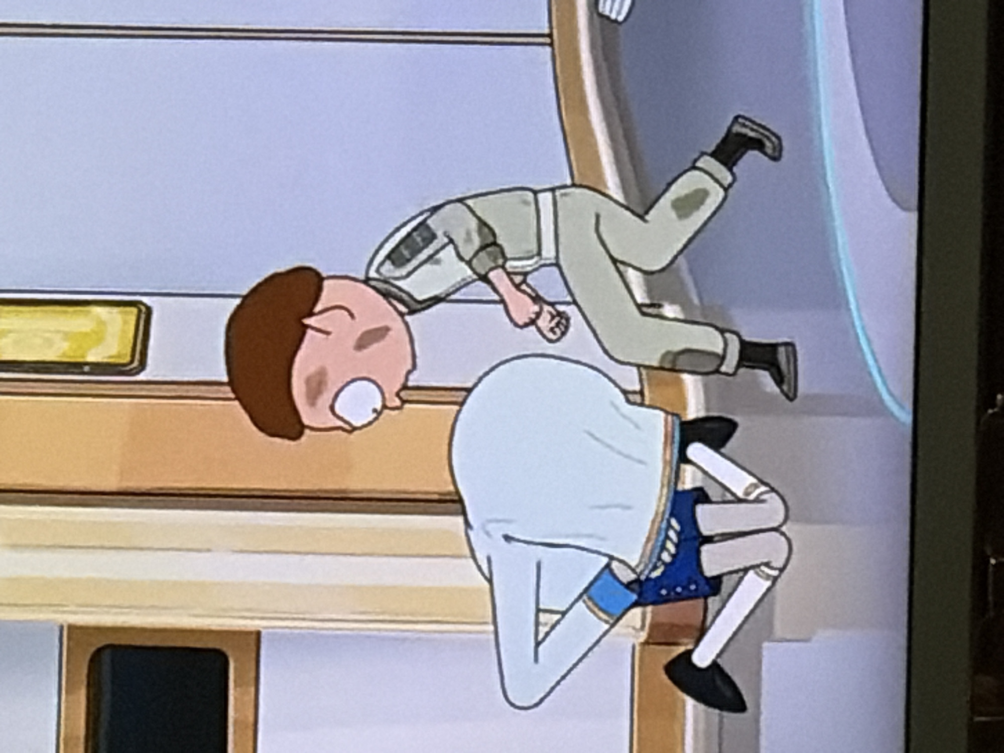 In an indoor environment, two animated characters are depicted. On the right is a male character with brown hair and pale skin, wearing a light green jumpsuit. He is in motion, with his right leg forward and his left arm extended, fist clenched. He appears to be looking to his left, towards the other character. On the left, a second character is bent over, with their head obscured by their body. This character is wearing a light blue long-sleeved shirt, dark blue shorts with decorative stripes, and white stockings with black shoes. Their posture suggests they are kneeling or bowing. The background features architectural elements with vertical cream and white stripes, and a dark rectangular window on the left. The scene is set against a light blue wall. The lighting is even and appears to be artificial.