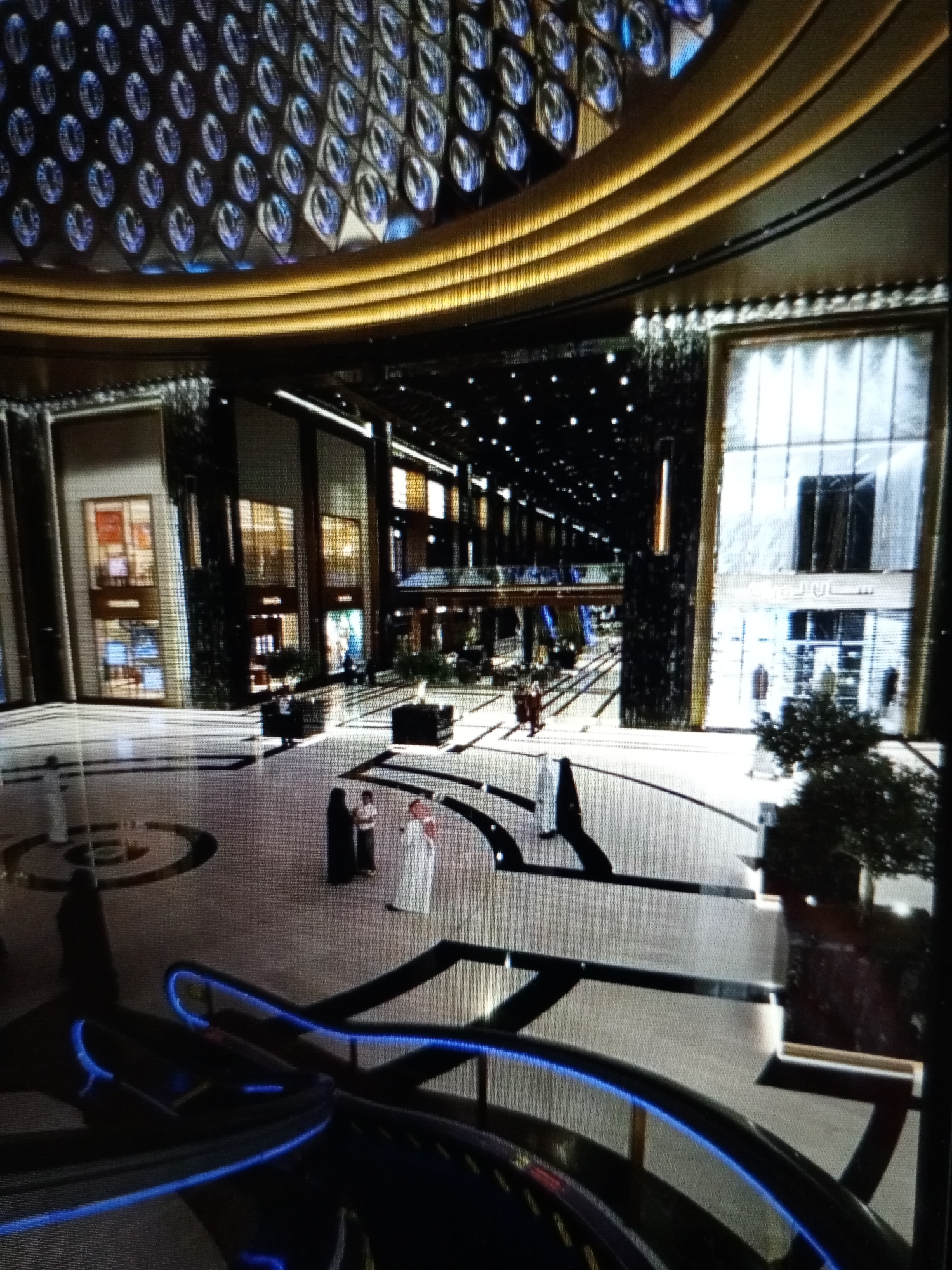 The image depicts an interior view of a large, modern shopping mall. The architecture features a grand, multi-level design with a prominent circular atrium. The ceiling is adorned with a decorative pattern of circular, textured elements, illuminated with blue light, and a golden, curved structure above. The mall's interior is spacious, with multiple levels visible, connected by escalators and walkways. The floor is a light-colored material with dark, geometric patterns.
Several individuals are present in the mall. In the foreground, a dark-colored escalator with blue lighting is visible. Further into the mall, on the ground level, three people are seen standing together, two in traditional Islamic attire (one in a black abaya and headscarf, the other in a white thawb and ghutra) and one person in lighter colored clothing. In the middle ground, several people are walking through the mall, some in groups. In the background, shops and store windows are visible, some with displays. The overall environment suggests a busy, upscale commercial space. There is no indication or possibility to determine the location in the city of Golbembal, Nigeria, as the image does not provide any identifying markers for that location.