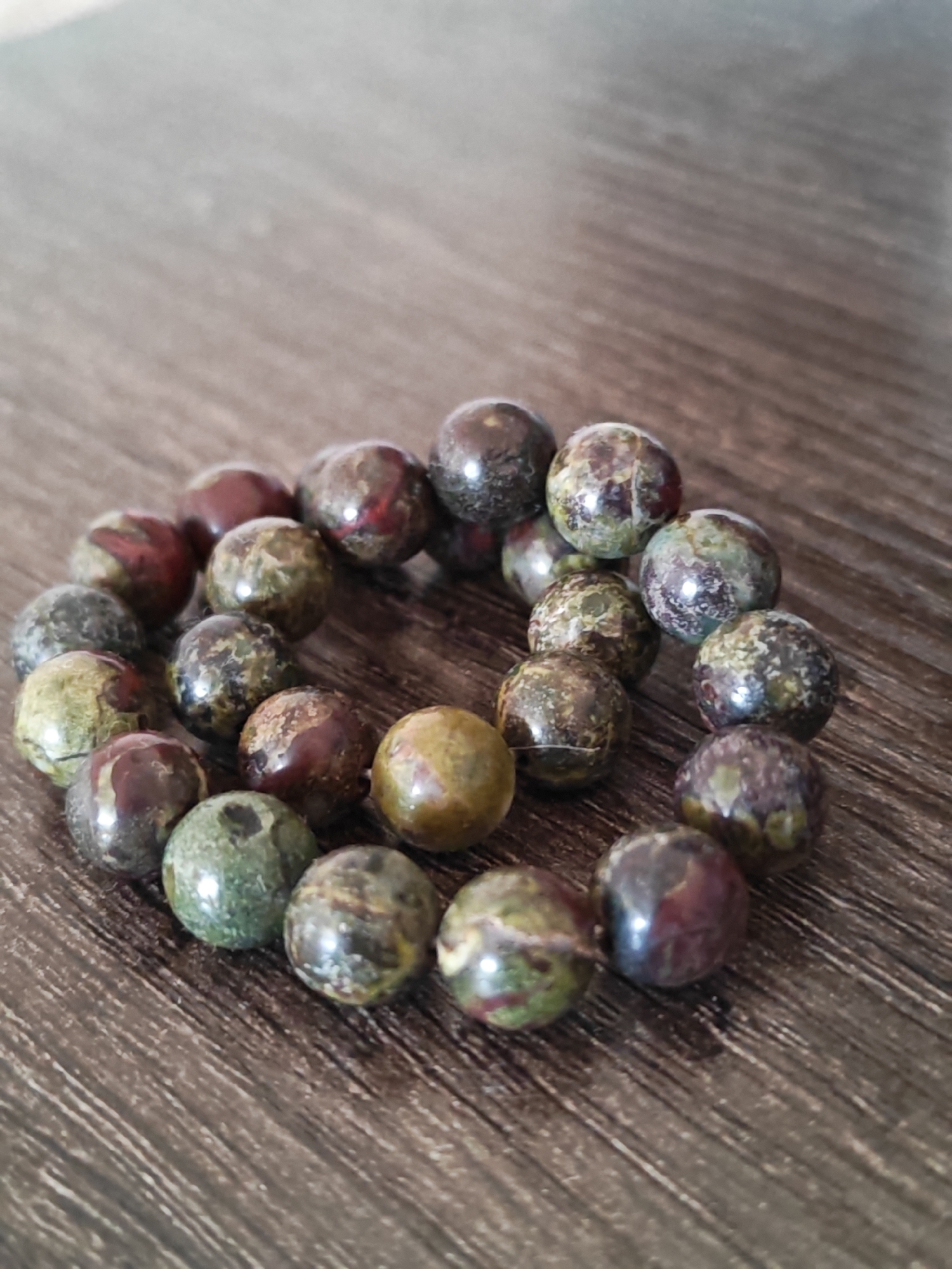 A bracelet made of round, polished stones with a variegated pattern of green, brown, and deep red hues is arranged in a circular shape on a dark wooden surface. The bracelet is the primary subject of the image, with the wooden surface serving as the background. The lighting creates a soft reflection on the stones. There are no people or other objects visible in the scene. The image does not provide any information that would allow for the determination of its location in Vanadzor, Armenia.