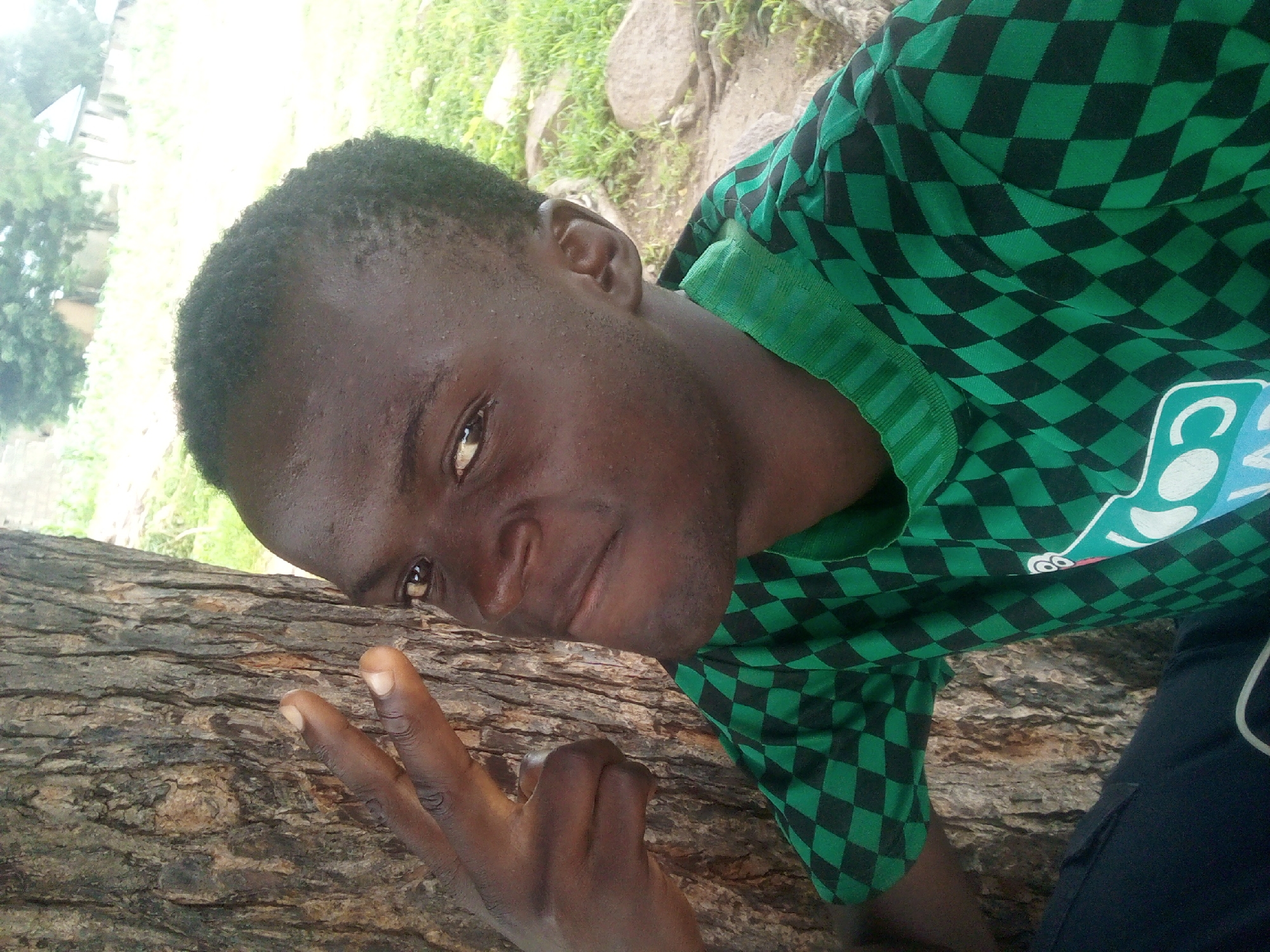 In an outdoor setting, a young Black male is pictured leaning against a tree trunk. He is wearing a green and black checkered t-shirt and dark pants. His right hand is raised, with his index and middle fingers extended in a peace sign. His gaze is directed towards the camera with a slight smirk. The background consists of greenery and rocks, suggesting a natural or semi-rural environment. The lighting indicates it is daytime.