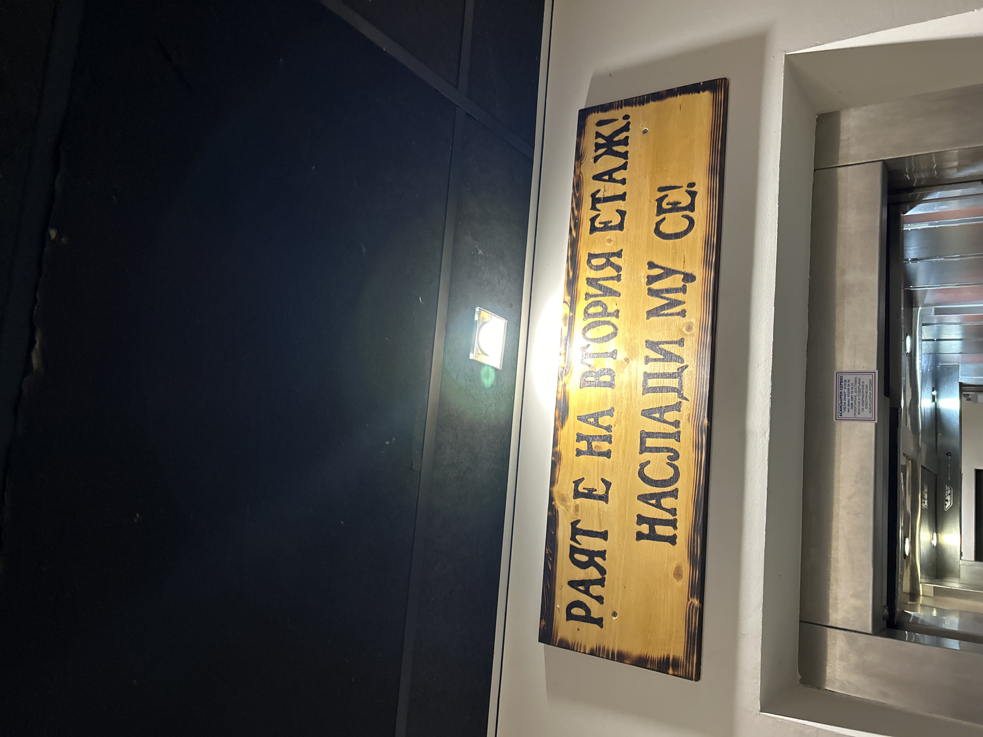 The image shows a sign in Bulgarian that reads "The heaven is on the second floor! Enjoy it!" The sign is made of wood and is placed near an elevator. The elevator appears to be made of stainless steel, and the building's walls are painted white. A small safety sign is attached to the elevator. The location is likely a building in Plovdiv, Bulgaria, based on the Bulgarian text.