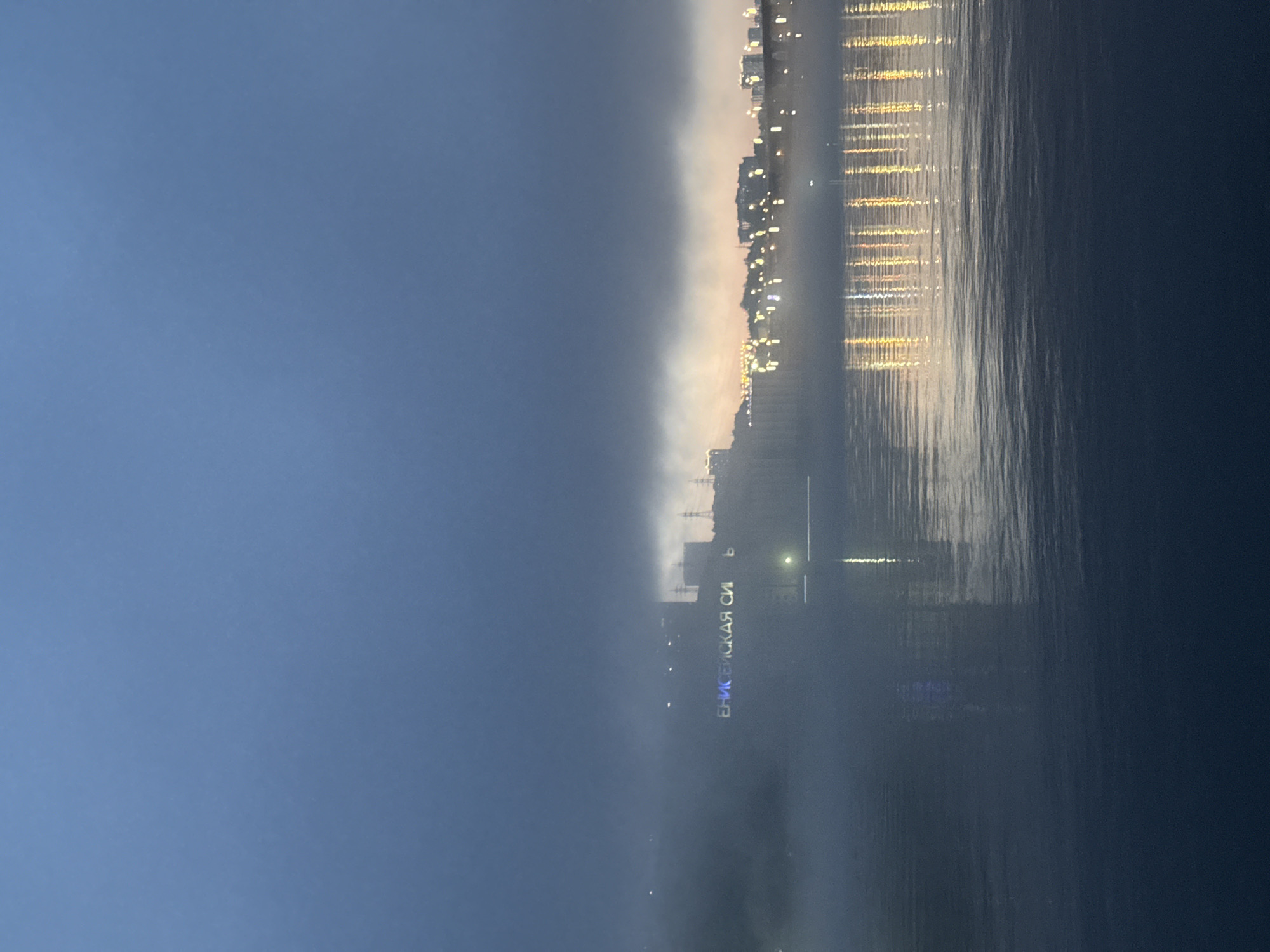 The image depicts a waterfront scene in Krasnoyarsk, Russia, at dusk or night. The Yenisey River is visible, with reflections of city lights. The skyline consists of buildings and infrastructure, illuminated by artificial lights. The sky is a gradient of blue, transitioning to a lighter hue near the horizon. The image is oriented sideways.