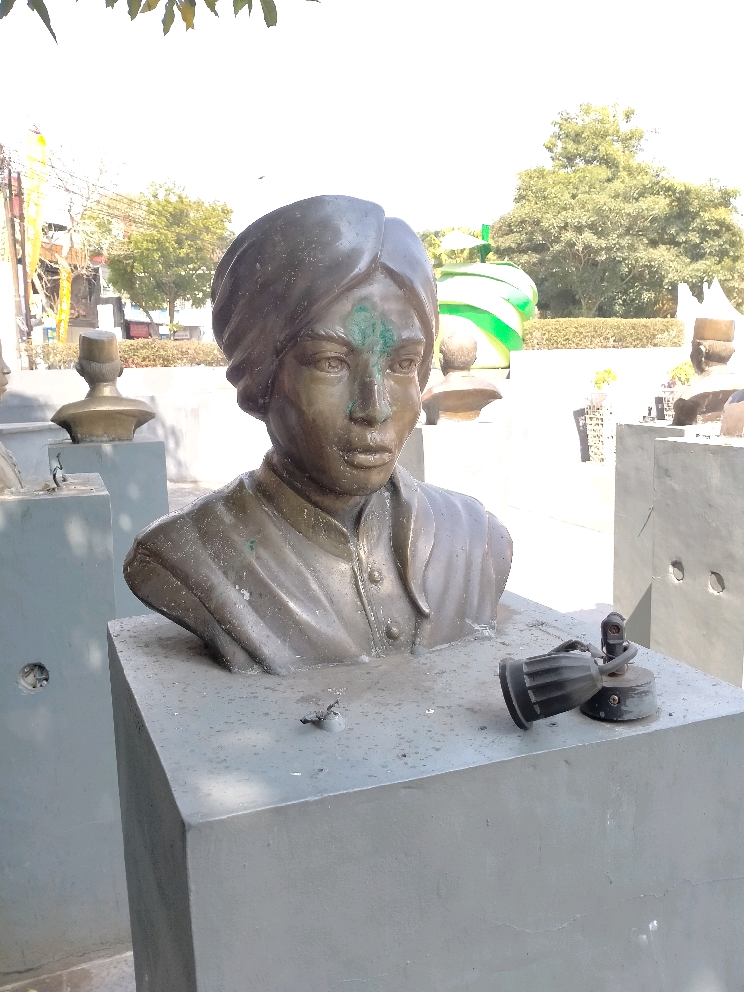 The image shows a close-up of a bronze bust of a person wearing a turban, positioned outdoors on a grey concrete pedestal. The bust is dark metal with some green patina. A small black object with a metal base and wire is attached to the pedestal to the right of the bust. In the background, other similar busts on pedestals are visible, along with trees, some buildings, and what appears to be a brightly colored green and white sculpture. The location is identified as the Tugu Pahlawan Batu in the city of Batu, Indonesia, which is a memorial to national heroes. The lighting suggests it is daytime, with bright sunlight and shadows.