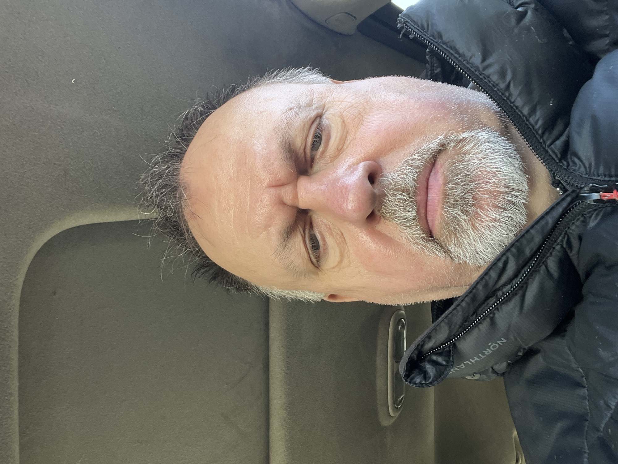 A close-up, slightly upward-angled selfie of a man with graying hair and a salt-and-pepper beard. He's wearing a black Northland jacket. The background shows a car interior in gray tones. The location is not evident in the image; therefore, it cannot be stated this photo was taken in Podolsk, Russia.