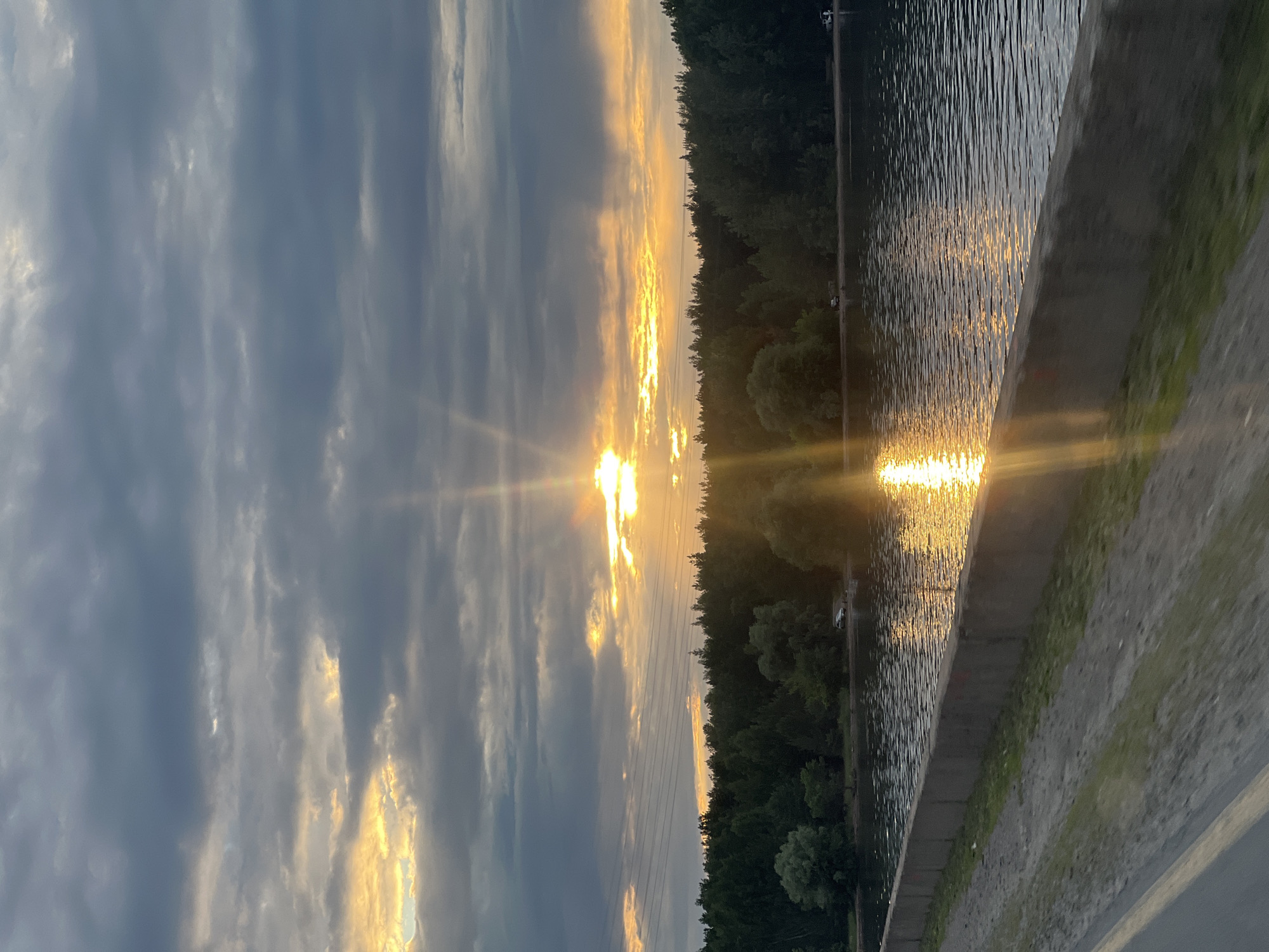 The image depicts a sunset over a body of water, likely a river or lake, in Noviy, Russia. The sky is filled with dramatic, dark gray clouds, with shafts of golden sunlight piercing through, creating a dramatic contrast. The sun is low on the horizon, casting a bright reflection on the water's surface. A dense line of dark green trees forms a backdrop along the far bank of the water. In the foreground, a concrete embankment separates the water from a dusty, gravelly path. The scene is serene and natural, with no visible people, though there are several vehicles parked along the tree line in the distance. The overall impression is one of natural beauty and tranquility.