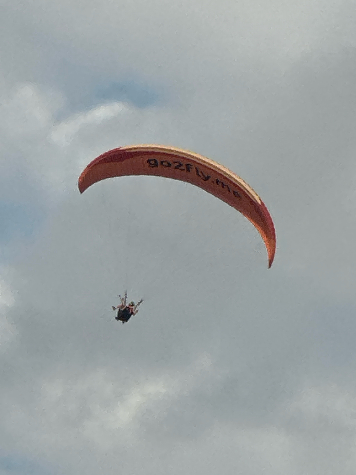 A paraglider with a pilot is suspended in the sky with a paraglider wing. The paraglider wing is an orange and yellow crescent shape with "gozfly.me" written on it in black letters. The pilot is seated in a harness attached to the wing. The paraglider is positioned in the upper half of the frame. The background is a cloudy sky. The image appears to have been taken from below. There is no indication of the location being Boreti, Montenegro within the image itself.