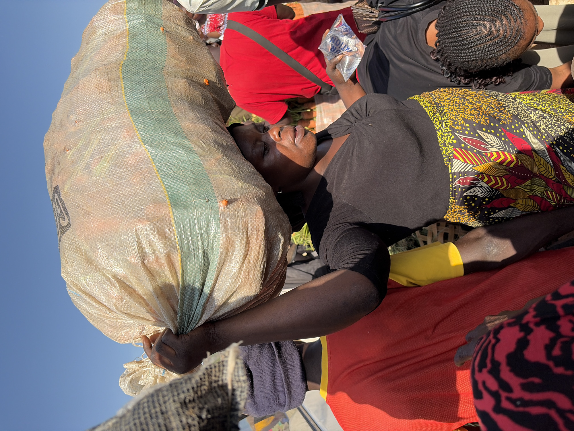 In Gura Top, Nigeria, a woman carries a large sack of produce on her head. She is dressed in a black shirt and a vibrantly patterned skirt. Another woman, wearing a red shirt, stands behind her, holding a clear plastic bag. In the foreground, a person in a red and black patterned garment is partially visible. The scene appears to be set outdoors, possibly at a market, with a clear blue sky overhead.