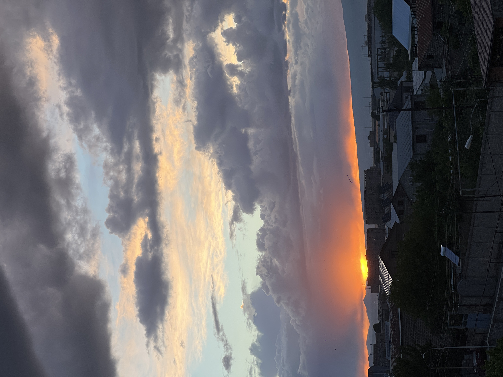 The image depicts a sunset over the city of Yerevan, Armenia. In the foreground are rooftops of buildings, with utility lines visible. In the midground, a vivid orange glow dominates the horizon, partially obscured by distant mountain silhouettes. The sky above is filled with a mixture of dark, heavy clouds and areas of lighter blue, some illuminated by the setting sun. The photograph appears to have been taken from an elevated position.