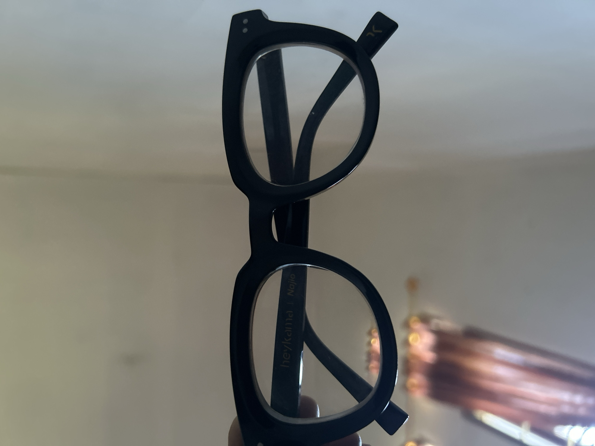 The image shows a pair of black-rimmed eyeglasses held up against a blurred background. The glasses have a round frame shape with clear lenses. The left arm of the glasses has the inscription "heyKama | Najio". The background appears to be an indoor setting, possibly a room with neutral-colored walls. There are blurred objects visible in the background, including what appears to be a curtain or drapery with brown and white vertical patterns. There is no indication in the image that suggests it was taken in the city of Lhokseumawe, Indonesia.