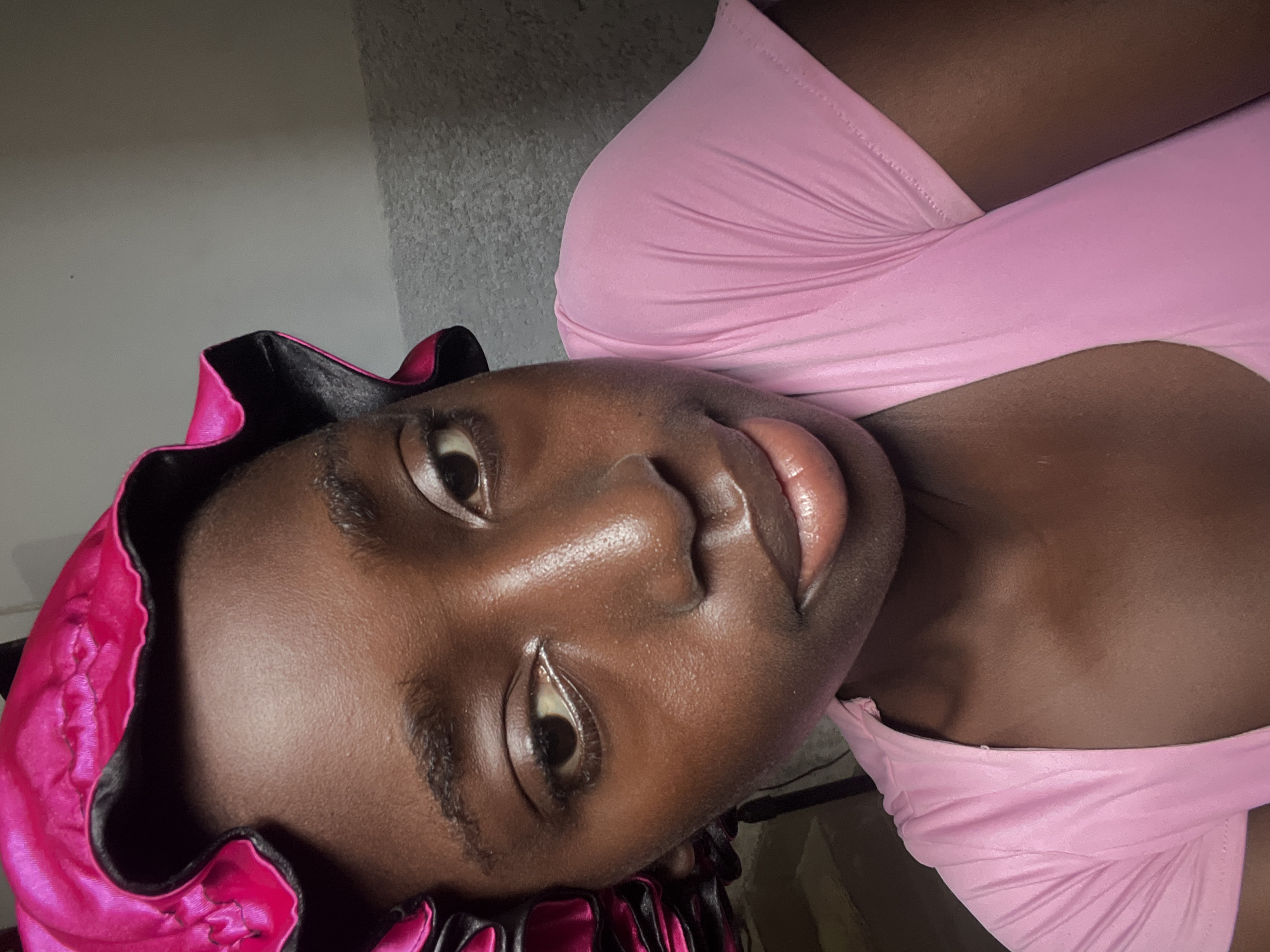 A close-up, slightly angled photograph captures a woman wearing a pink satin bonnet and a pink top. The background appears to be a wall with a textured finish. The location is not discernible from the image.