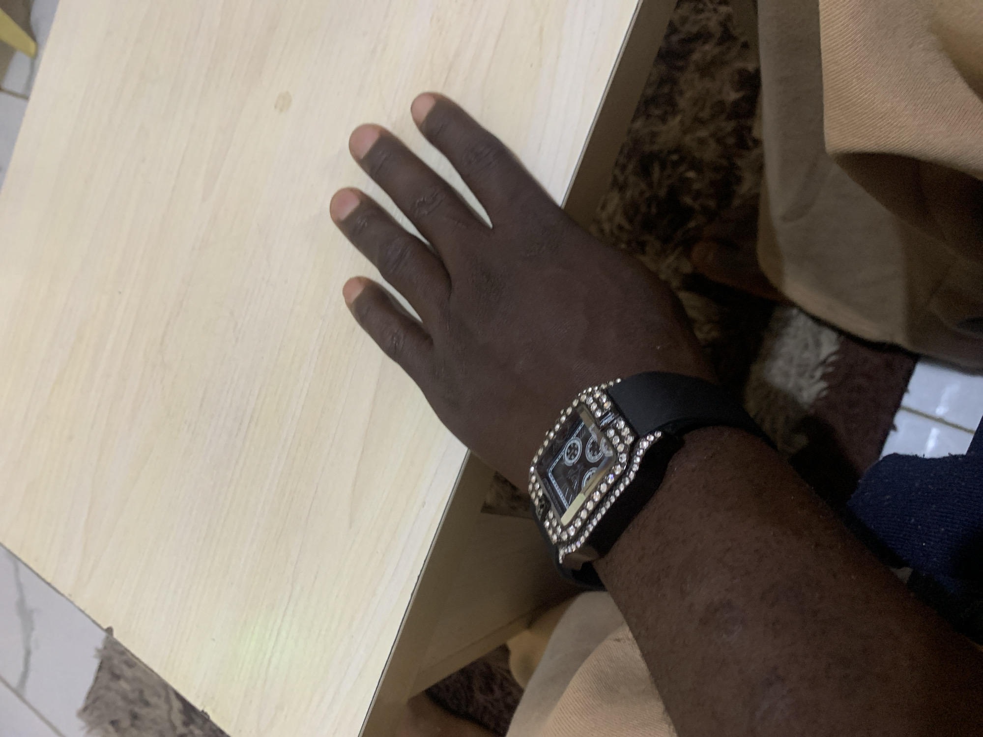 A close-up, overhead shot shows a person's right hand resting on a light-colored wooden table. The hand is dark-skinned, and the fingers are slightly spread. A black wristwatch with a jeweled bezel is worn on the wrist. The background includes parts of a patterned rug and possibly some tiled flooring. The image appears to be taken indoors. The location of Jos, Nigeria, is not discernible from the visual information provided in the image.