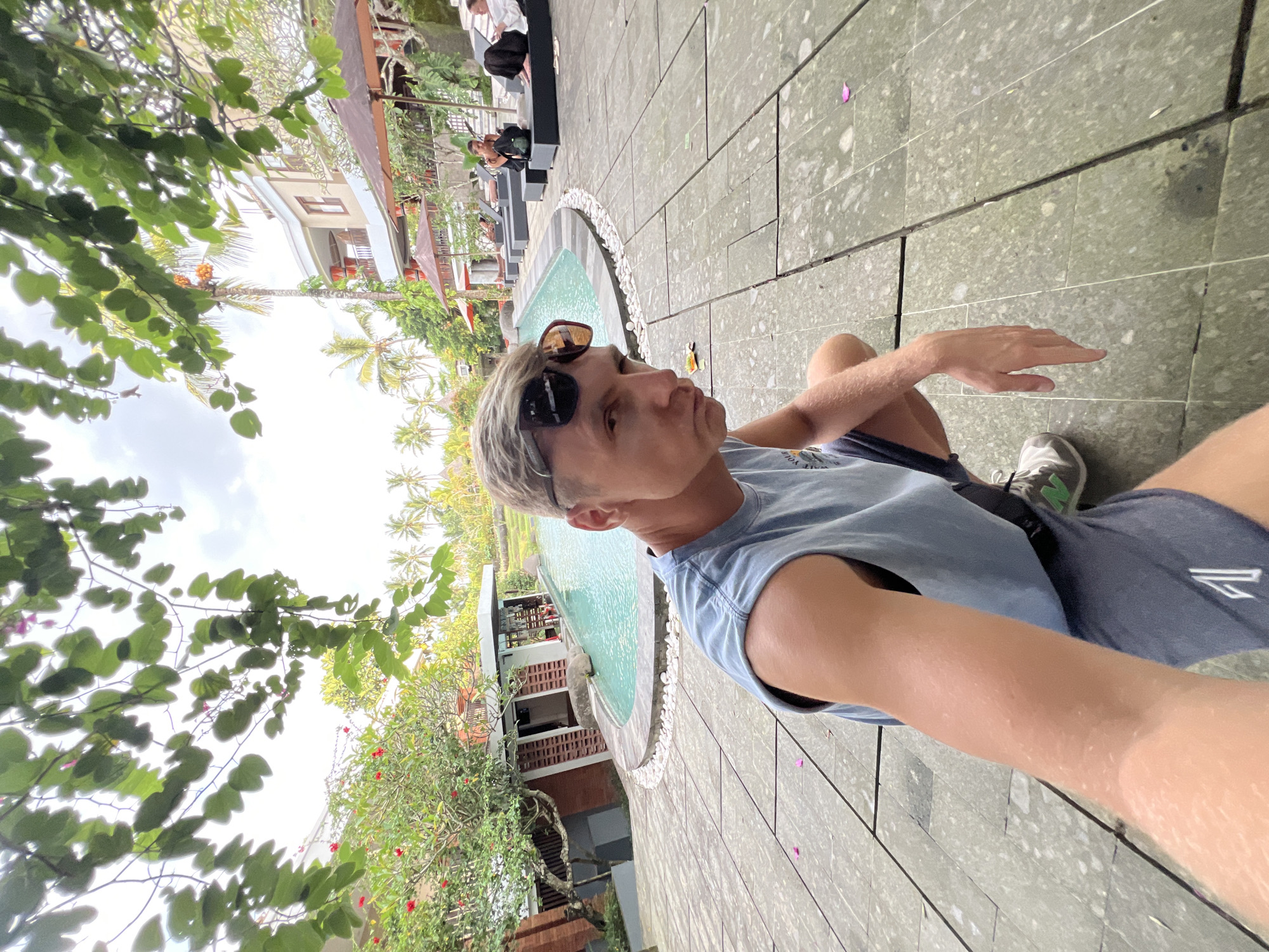 A man with sunglasses and a sleeveless shirt is sitting on a tiled surface next to a pool. The location is the Ubud District, Indonesia. The surroundings include buildings, palm trees, and other vegetation.