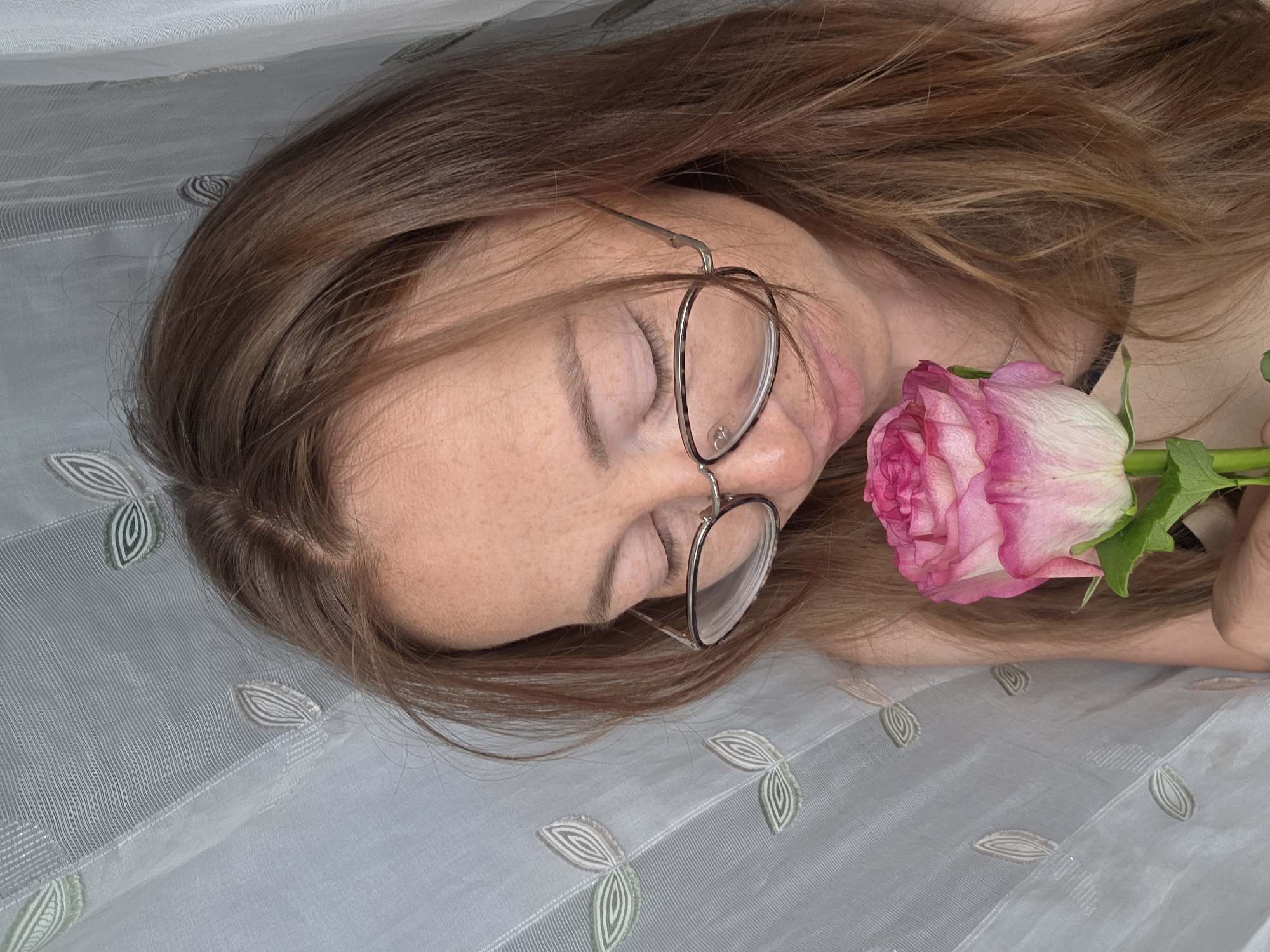 The image shows a person lying on a light gray fabric with a leaf pattern. The person has long, light brown hair and wears glasses. A pink and white rose rests near their face. The location is unknown and cannot be determined from the image.