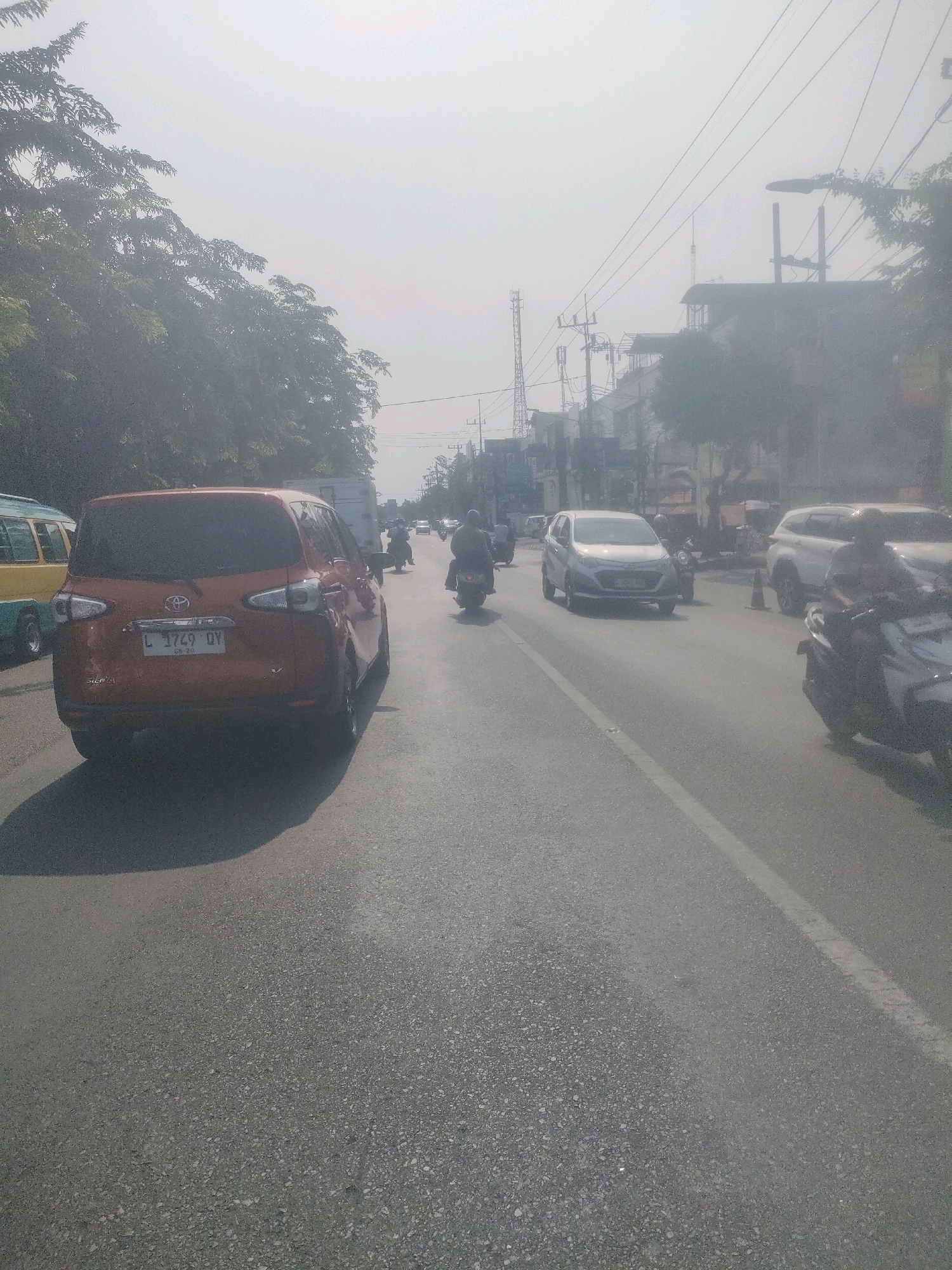 This is a photograph taken on a hazy, sunny day on a street in Surabaya, Indonesia. The viewpoint is from a vehicle moving along the right lane of a two-lane road. In the foreground, a red Toyota Sienta car with license plate L 1749 DY is visible. To the left, a yellow and blue bus is partially visible. Several motorcycles are also present on the road, some carrying riders. In the background, there are buildings and utility poles, characteristic of an urban environment. The road surface is asphalt. The haze may indicate air pollution or atmospheric conditions common in the region.