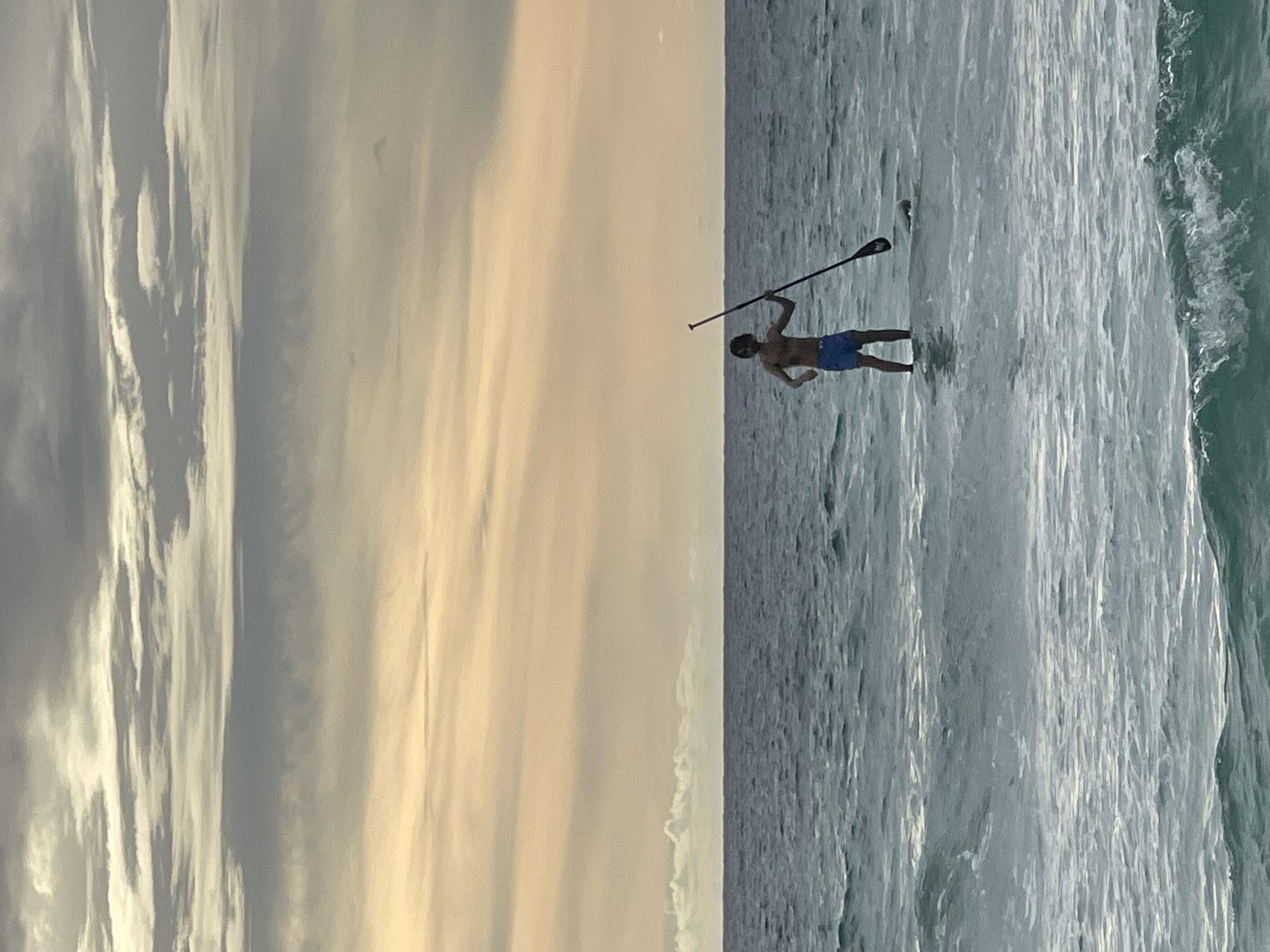 A full shot shows a young man paddleboarding in the ocean at Karon Beach, Thailand. The man, wearing blue swim trunks, stands on the paddleboard, holding a paddle. The water is a deep blue with gentle waves, and the sky above is filled with dramatic, layered clouds that are tinged with yellow and orange from the setting sun. The horizon line is sharp, separating the water from the sky.
