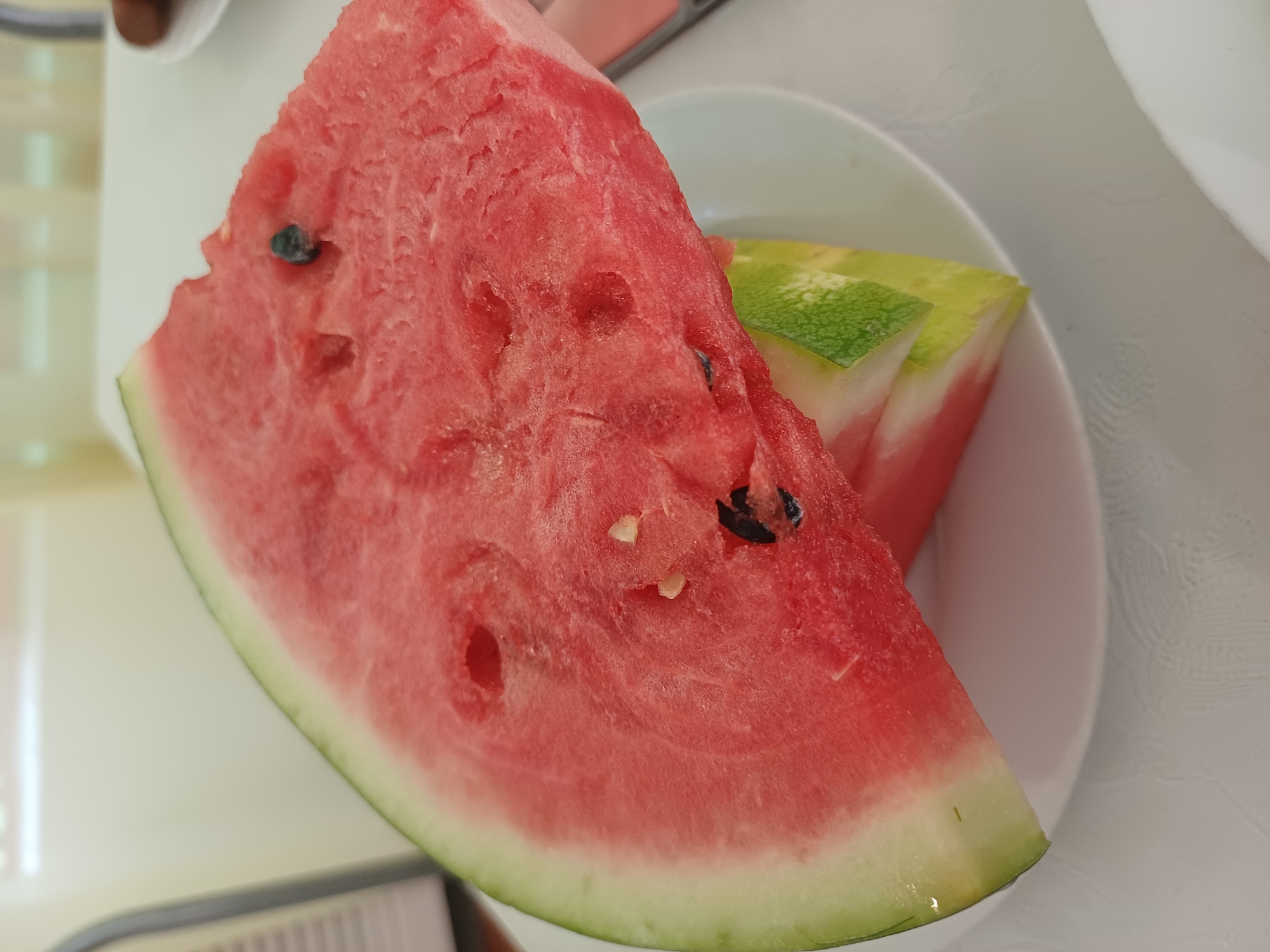 A close-up shot displays two slices of watermelon on a white plate. The watermelon slices are triangular, with the flesh being a vibrant red and the rind a light green. Several black seeds are visible within the flesh of the top slice. The background is blurred, suggesting an indoor setting with soft, natural light, possibly a restaurant or dining area. The image is taken from a slightly elevated angle. There is no indication of location in the city of Varna, Bulgaria.