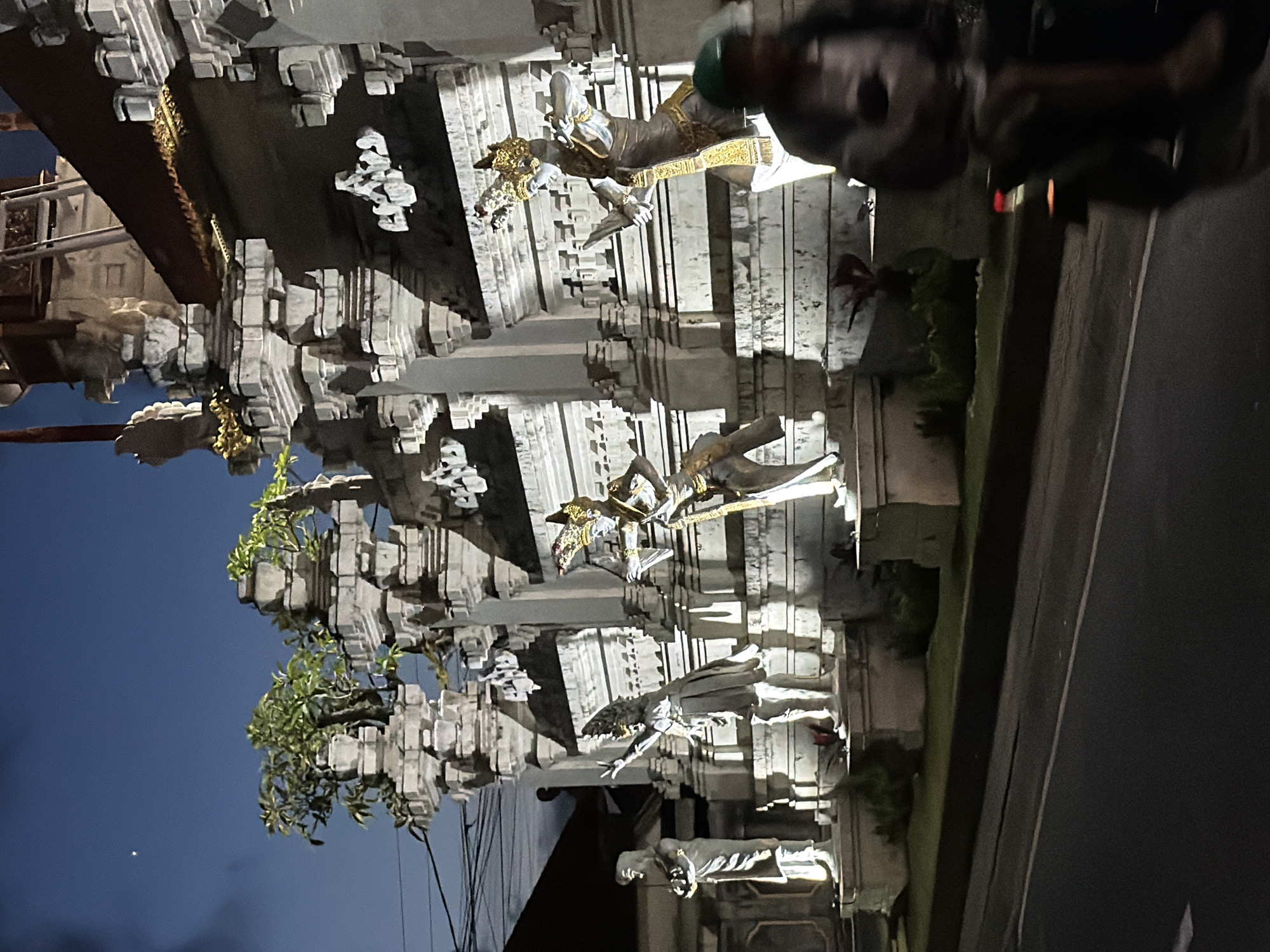 Captured at night in Kutuh, Indonesia, the photograph showcases a Balinese temple gateway. The architectural structure features intricate carvings of deities, statues, and traditional Balinese motifs. The gate is built from light-colored stone. Lush greenery and plants accent the architecture. A road with vehicular traffic is visible alongside the temple. A person is near the gate. The night sky serves as a backdrop. Electrical wires are near the structure.