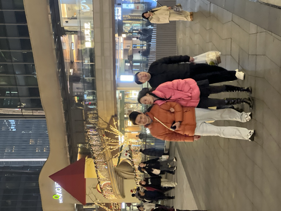 The image depicts an urban pedestrian area at night outside The King Glory Plaza in Liangjiang New Area, Chongqing, China. In the foreground, a group of three East Asian individuals walks left-to-right across a grey geometric-tiled pavement. The leading individual, a male with a medium build, wears an orange puffer jacket, light grey pants, and white sneakers, holding a dark object in his left hand while his right arm is linked with the woman to his right. This woman, of slender build, wears a pink puffer jacket, a black skirt, black tights, and black knee-high boots, looking towards the man on her left. To her right, a second male, of heavier build, wears a black top, black pants, and white sneakers, carrying a translucent plastic bag with contents in his left hand. Numerous other pedestrians are visible in the background, walking in various directions. On the far right, a solitary East Asian female, dressed in a light beige padded jacket and matching pants, stands still, holding a small bag. The background features a multi-level commercial building identified by the illuminated "THE KING" logo and Chinese characters. A red conical roof structure, resembling a turret, is visible, connected to a curved pedestrian ramp adorned with numerous string lights. Trees also decorated with string lights are present. Modern high-rise buildings with illuminated windows are discernible in the distant background. Artificial lighting sources illuminate the entire scene.