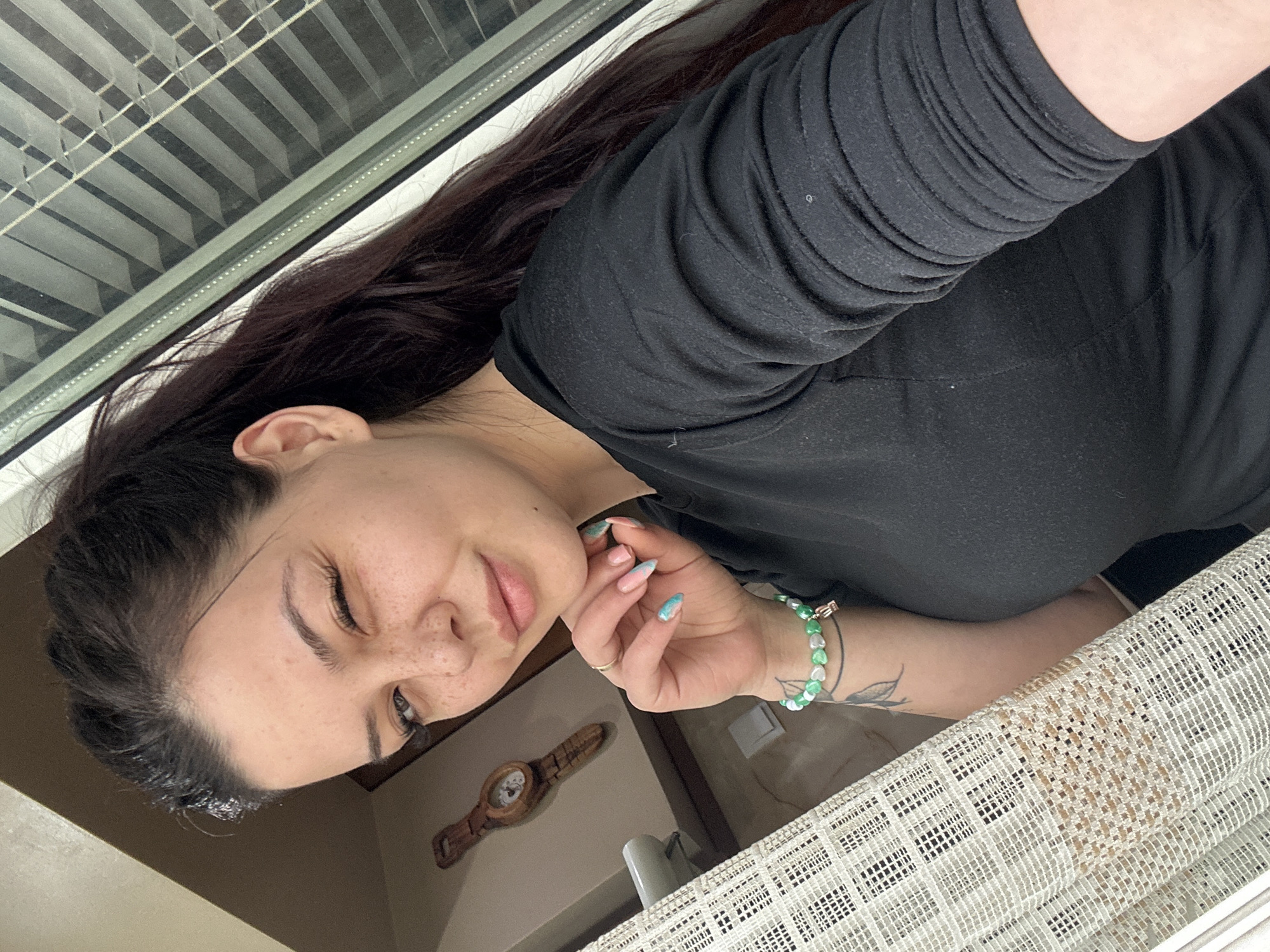 A woman with long dark hair and fair skin is indoors, possibly in Sliven, Bulgaria, though location is unconfirmed. She wears a black long-sleeved shirt and a green and white beaded bracelet on her left wrist, which also has a small tattoo. Her left hand is near her face. The background includes a window with closed blinds, a wall with a decorative wooden clock, and a white, patterned fabric. The angle of the image suggests it was taken from above, looking down at the woman.