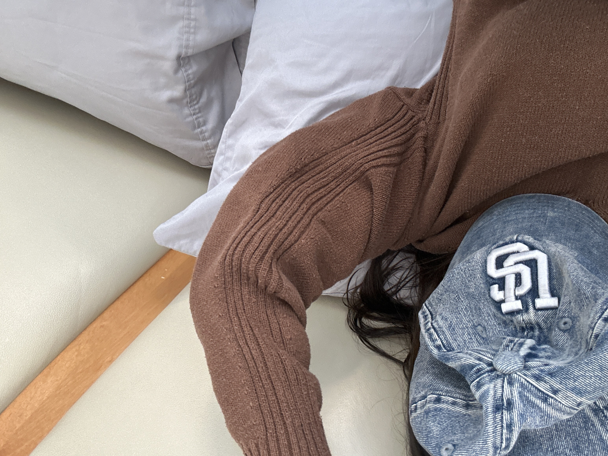 A top-down shot reveals a person resting on a bed, with a brown knit sweater covering their upper body. A blue denim baseball cap featuring a white "SD" logo rests next to their head, partly covering their dark hair. The bed includes white pillows and a light-colored flat surface near the edge of the mattress. The location and specific city are not identifiable from the image.
