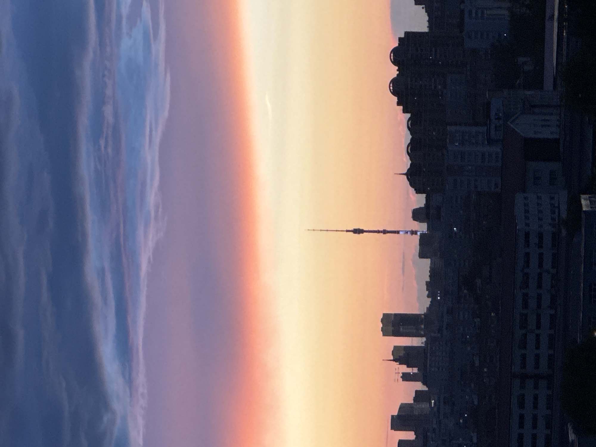 This is a vertical, medium-shot, silhouette photograph of a city skyline at dusk. The sky is a dramatic gradient of colors, transitioning from deep blue and purple at the top to vibrant pink and orange, and finally to a pale yellow near the horizon. Clouds are visible throughout the sky, catching the twilight colors.
Dominating the mid-ground is the Ostankino Tower, a prominent telecommunications tower in Moscow, Russia, lit with blue and white lights. To the left of the tower, a cluster of skyscrapers forms a more jagged silhouette against the colorful sky. To the right of the tower, a row of apartment buildings and other structures create a more uniform, dark silhouette. The foreground consists of a dark, dense urban landscape with multiple layers of buildings, including what appears to be a long, low-rise building with multiple windows. The overall impression is one of a city at the cusp of night, with the fading light casting long shadows and highlighting the architectural shapes of the urban environment. There are no people or animals visible in the frame.