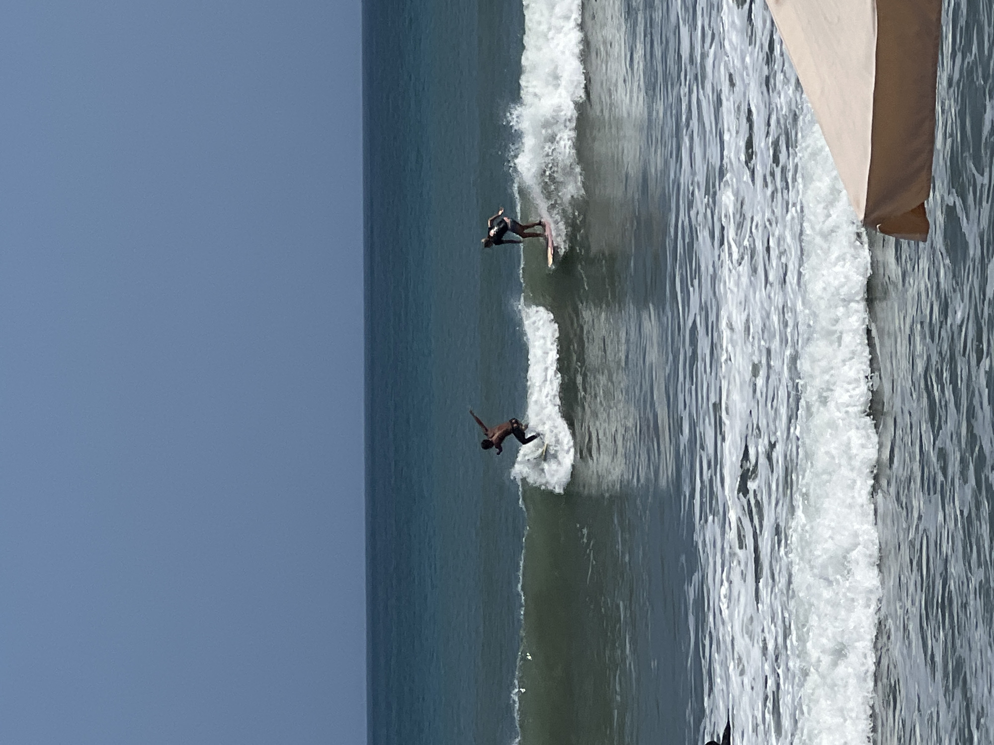 Two surfers are riding waves in the ocean. The surfer on the left is a dark-skinned male wearing dark shorts, riding a white surfboard. The surfer on the right is a light-skinned male wearing dark shorts and a dark top, riding a light-colored surfboard. The ocean is a deep blue-green with white foam from the waves. The sky is clear and pale blue. In the foreground, a tan umbrella is visible, partially obscuring the water. This scene is characteristic of Kuta Beach in Kuta, Indonesia, a popular spot for surfing.