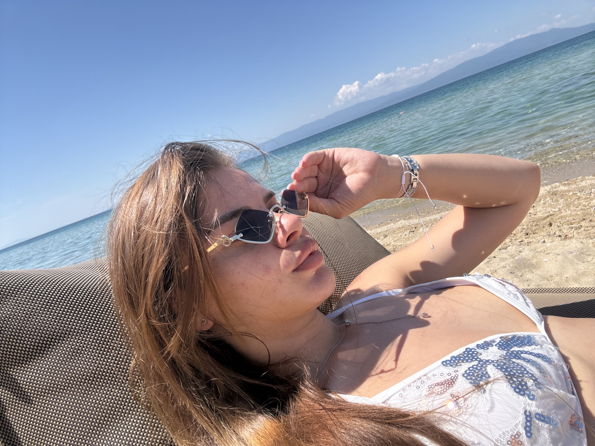 A woman is lying on a beach chair, facing away from the camera. The woman has long brown hair and is wearing a white bikini with silver sequins. She is also wearing sunglasses with gold frames and several bracelets on her right wrist. Her right hand is holding the sunglasses. A silver necklace is around her neck. The beach chair has a woven texture. In the background is a blue ocean and a clear blue sky. Hills are visible in the distance. The location is a beach in the city of Unknown, Unknown.