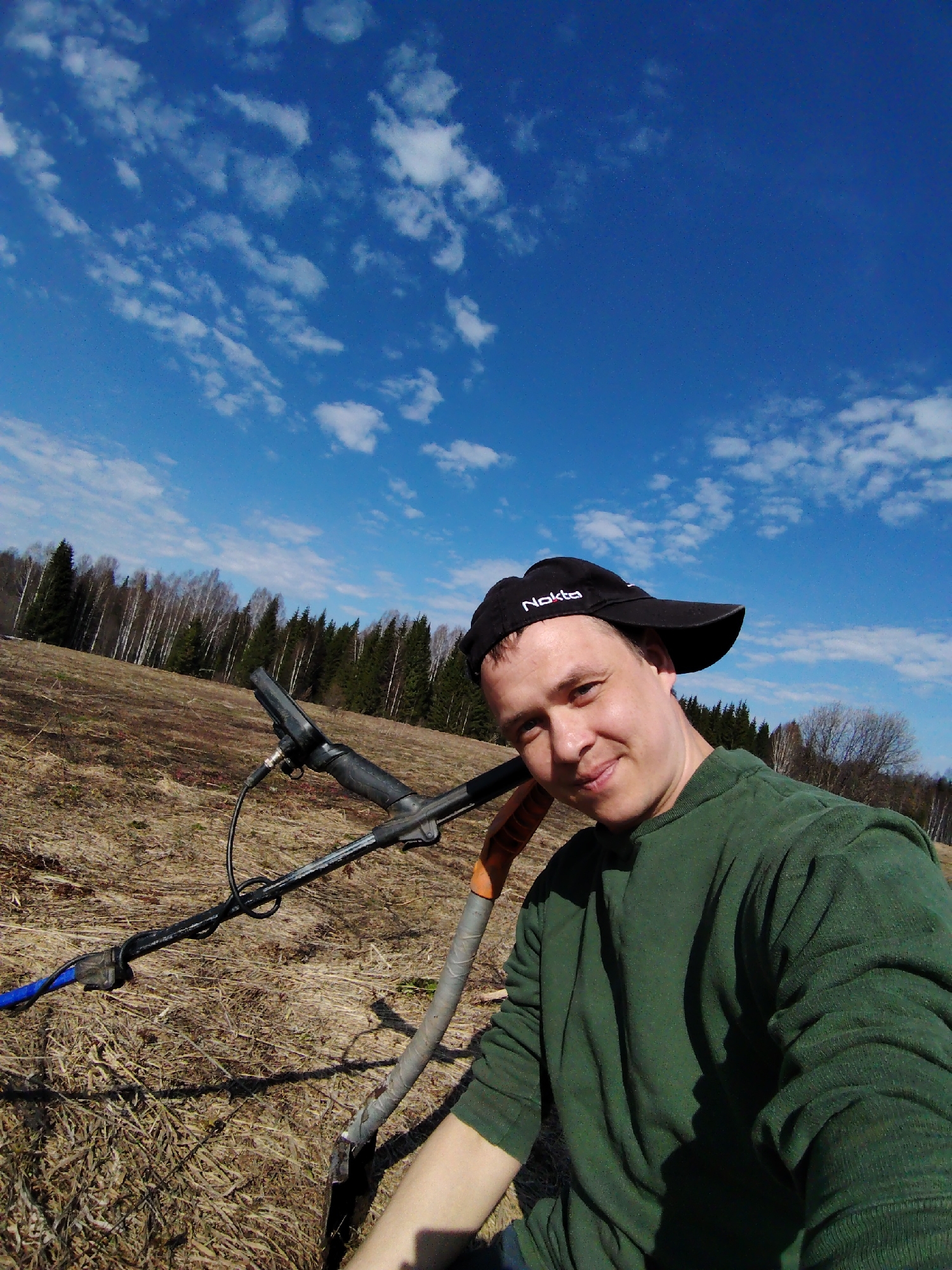 A man is captured in a selfie-style shot outdoors in an open field under a bright, partly cloudy blue sky, likely in Markovskii, Russia. He is fair-skinned, appears to be in his 30s or 40s, and is looking directly at the camera with a slight, friendly smile. He wears a dark green long-sleeved shirt and a black baseball cap with the word "Nokta" visible in white text on the front.
To his left, a black and grey metal detector rests on the dry, brownish grass, with its coil and a wire wrapped around the shaft clearly visible. Part of a shovel with a silver blade and an orange handle is also seen near the bottom right, suggesting he is engaged in metal detecting. The field is covered with sparse, dry vegetation, indicating a season like early spring after snowmelt or late autumn. In the background, a line of trees marks the edge of the field, featuring a mix of light-barked deciduous trees (possibly birch) and darker green coniferous trees.
The scene is brightly lit by the sun, suggesting daytime, likely late morning or early afternoon, with clear weather conditions. The man's expression suggests contentment, possibly enjoying his outdoor activity.