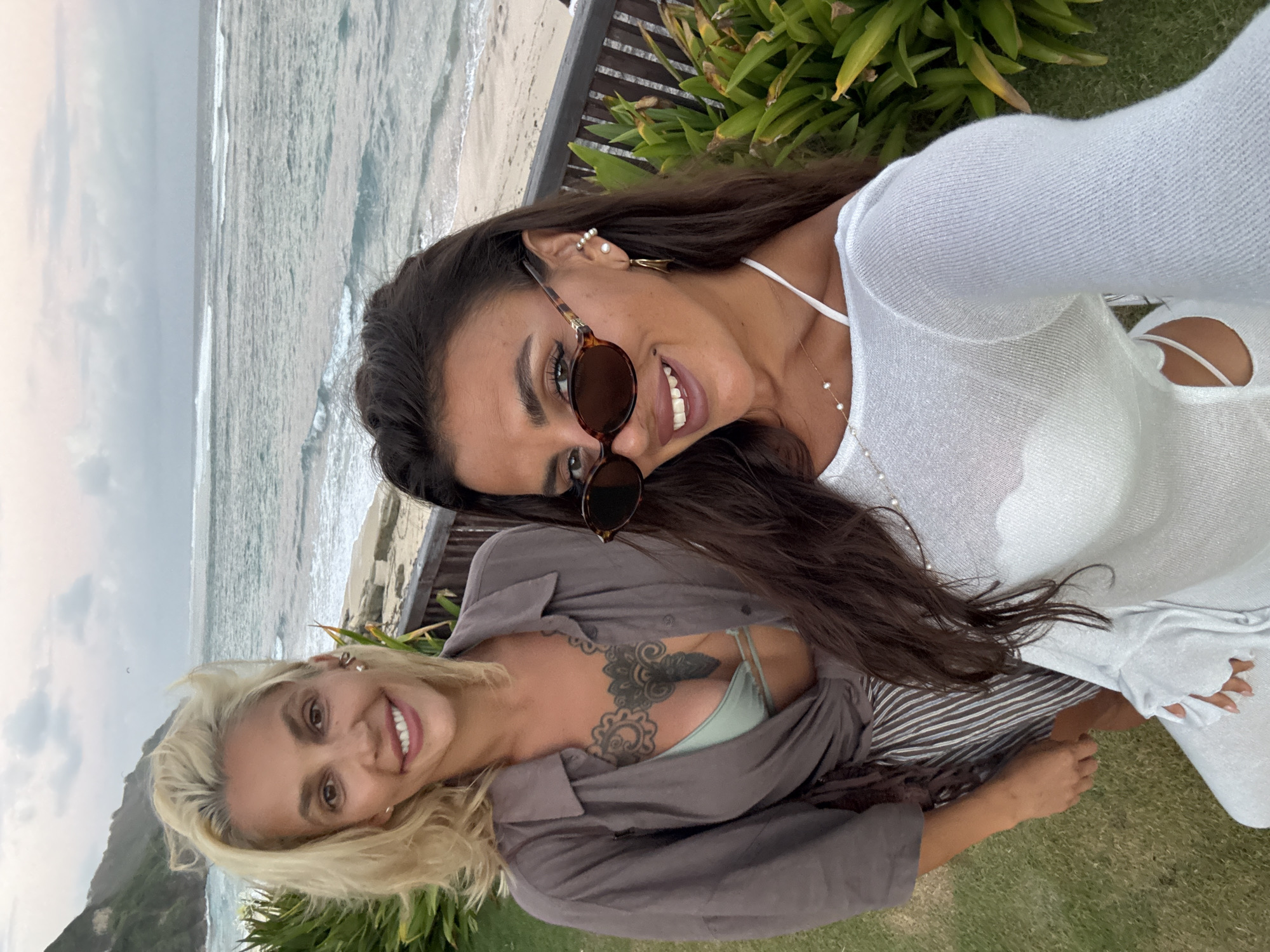 Two women are taking a selfie on a grassy area near a beach with the ocean in the background. The woman on the left has blonde hair and is wearing a grey shirt over a green bikini top and striped skirt. She has a large tattoo on her chest. The woman on the right has dark hair and is wearing a white top and sunglasses. They are both smiling. The location appears to be Kuta Selatan, Indonesia, given the distinctive coastline and vegetation. The sky is overcast, and the waves are visible in the ocean. There is a wooden railing and lush green plants in the foreground.