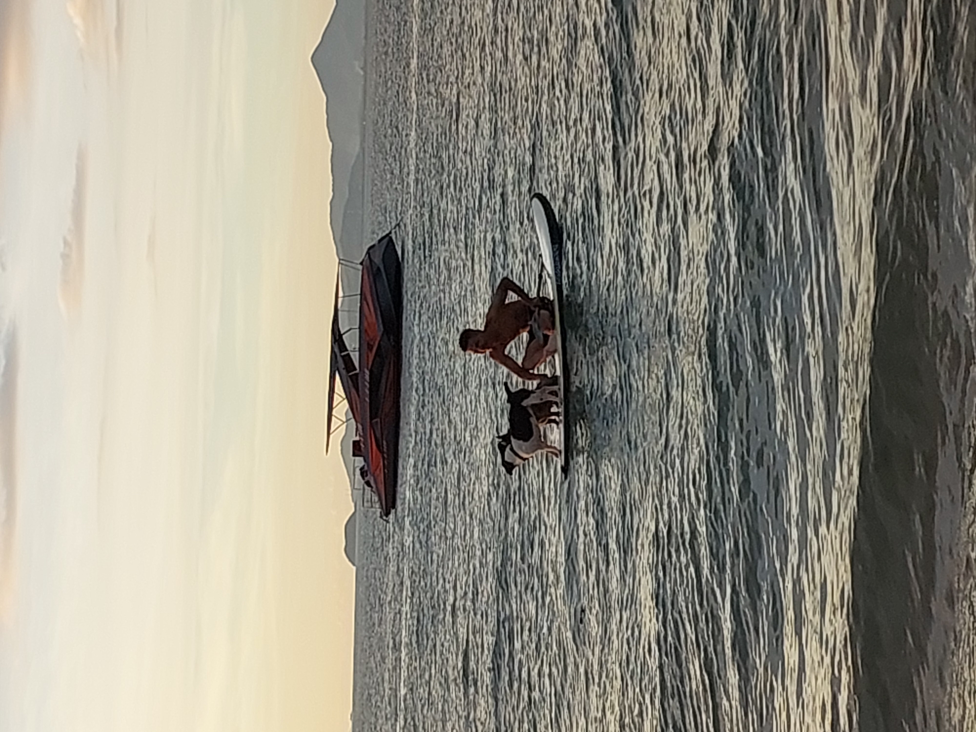 A man is sitting on a paddleboard in the ocean with two dogs. The man is shirtless and wearing shorts, and he is facing to the left. One dog is standing on the paddleboard, and the other is sitting next to it. The dogs are black and white. In the background, there is a red and black boat and land in the distance. The sky is cloudy and a warm color, indicating it is either sunrise or sunset. The ocean is wavy and dark. The location is Baan Mae Nam, Thailand.